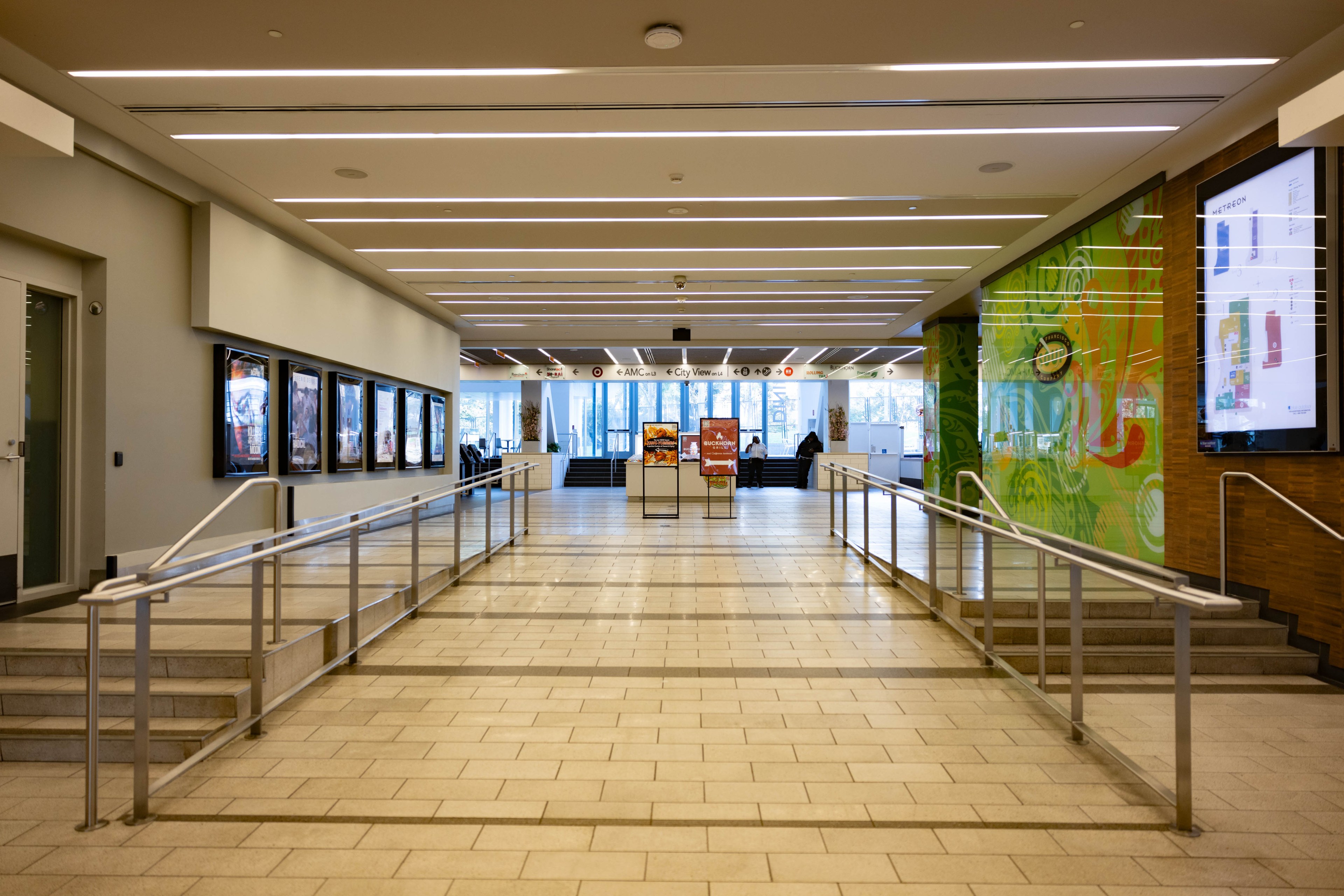A spacious hallway with a tiled floor, accented by railings on either side, leading to a sunlit area with signs and large windows. Walls display colorful posters.
