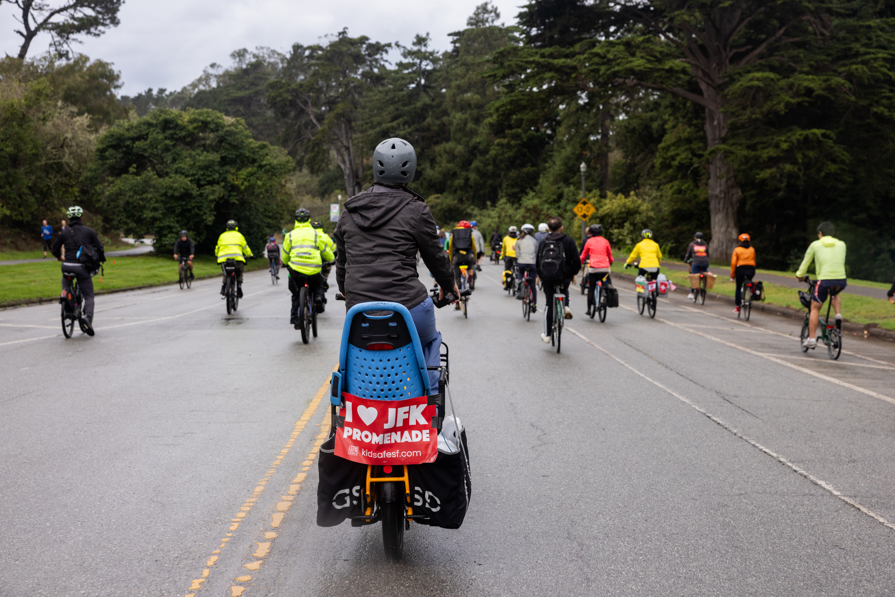 A group of people ride their bike through Golden Gate Park and on the back of one of the bikes hangs a sign that reads, &quot;I Love JFK Promenade.