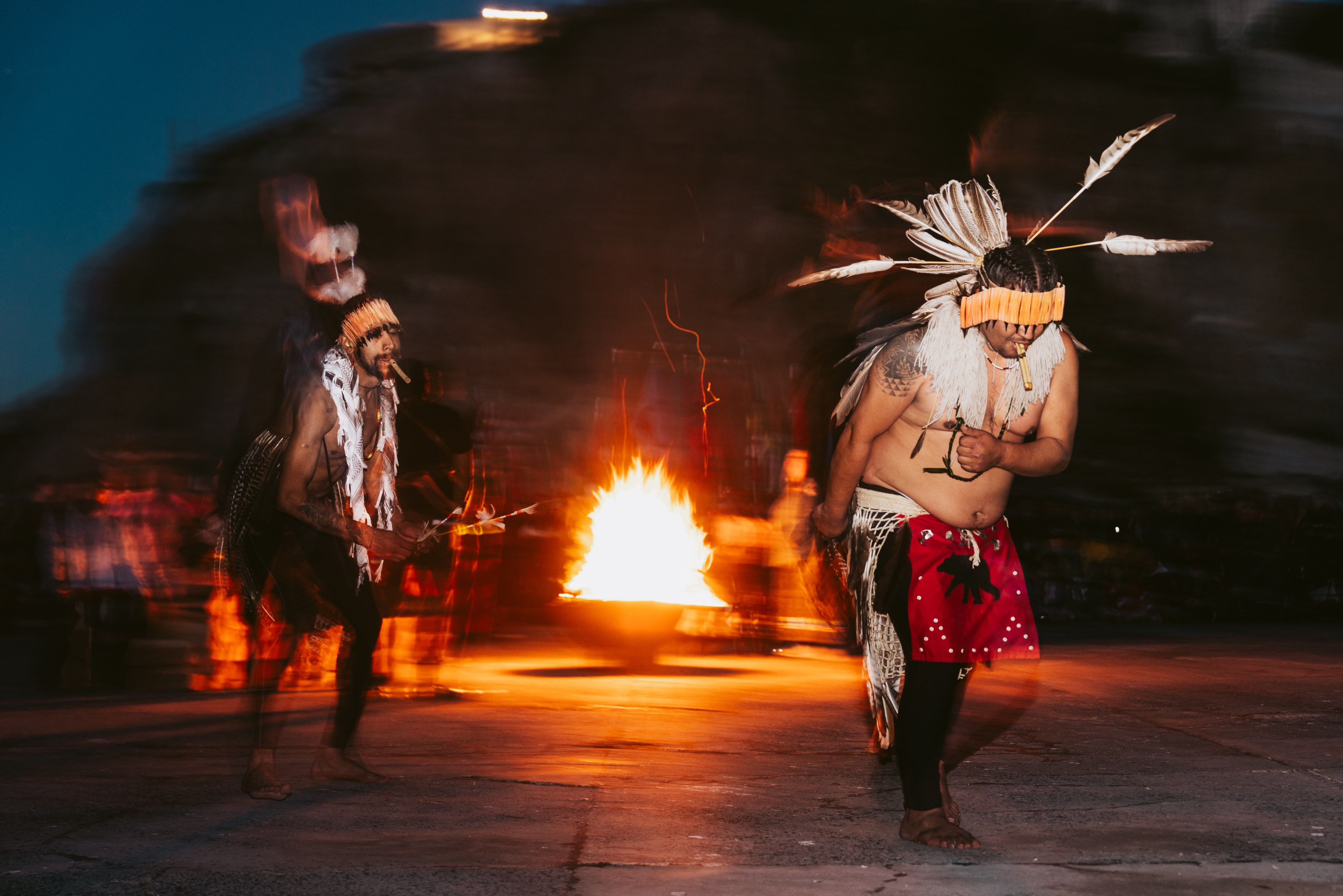 Two dancers in feathered headdresses dance by a fire under a dark sky.