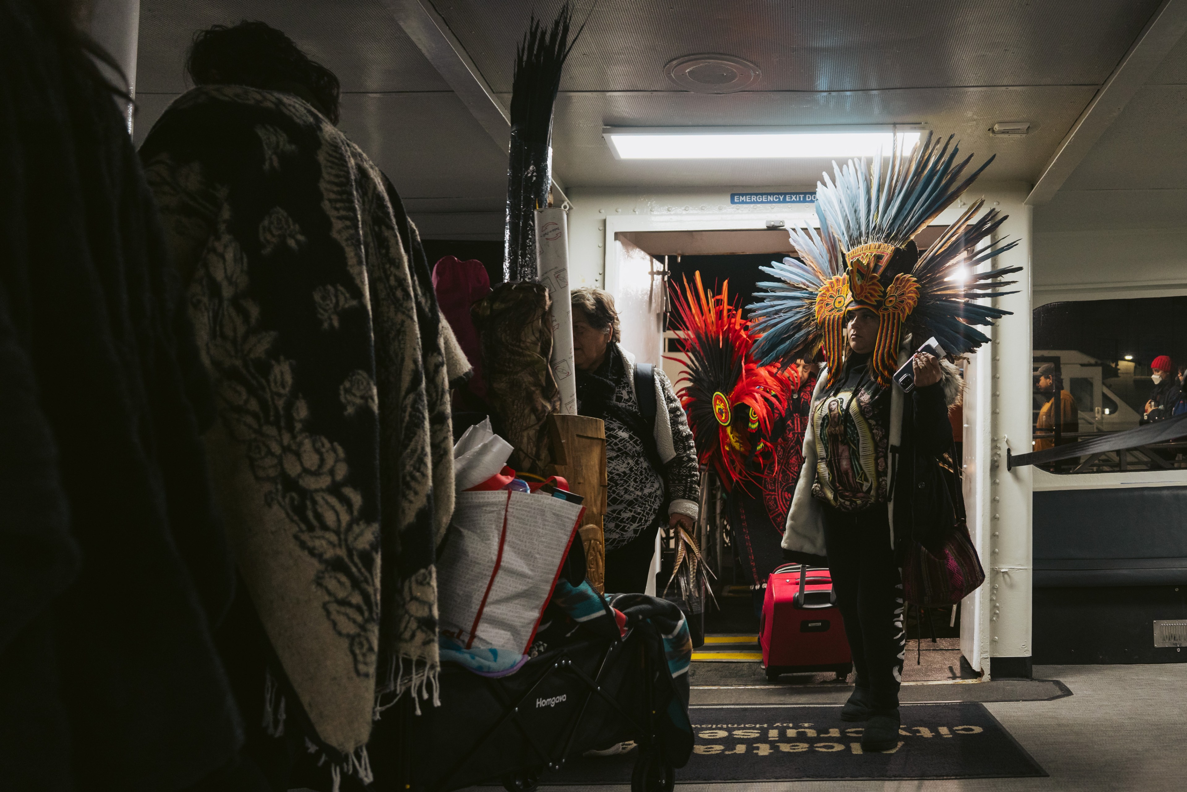 People adorned in indigenous regalia walk into the cabin of a ferry boat.