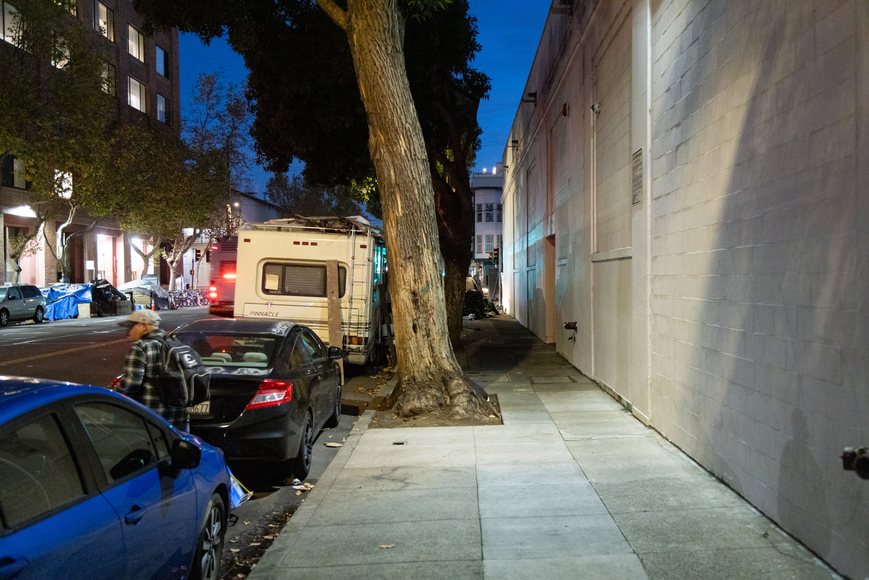 A row of cars with an RV are parked a brightly light side walk at night. 