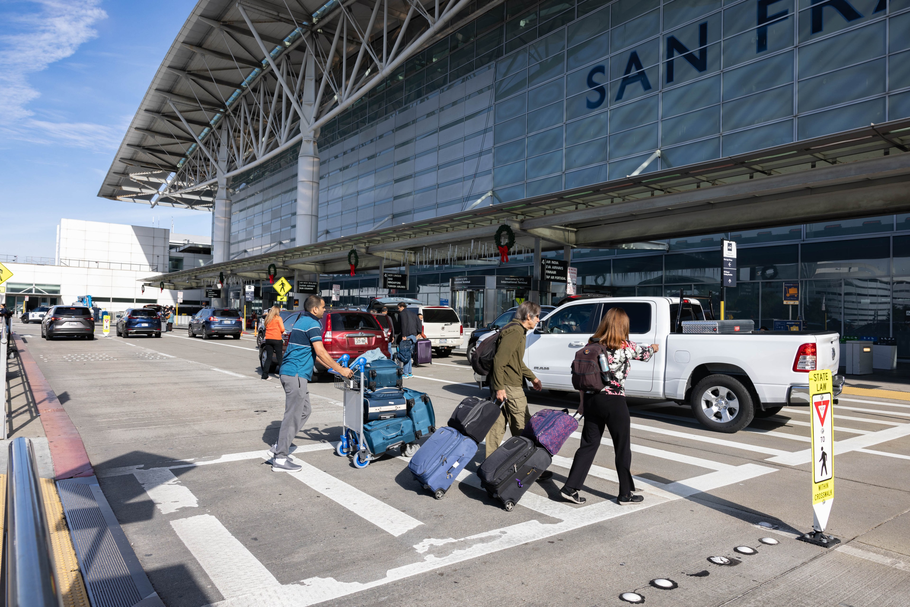 Travelers haul their luggage on a crosswalk outside an airport terminal.