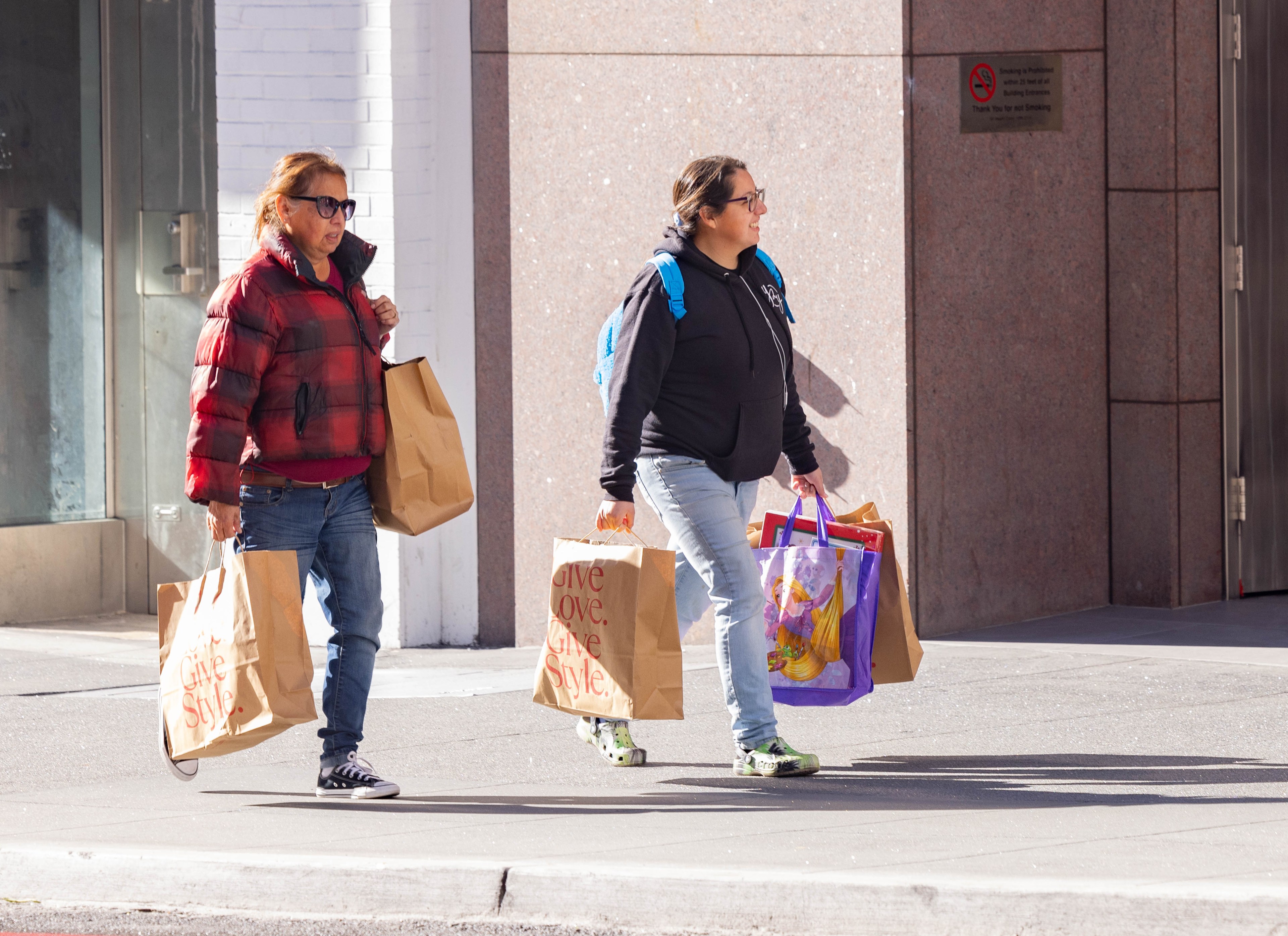 Two people walking on a sidewalk, carrying multiple shopping bags, with a sunny city backdrop.