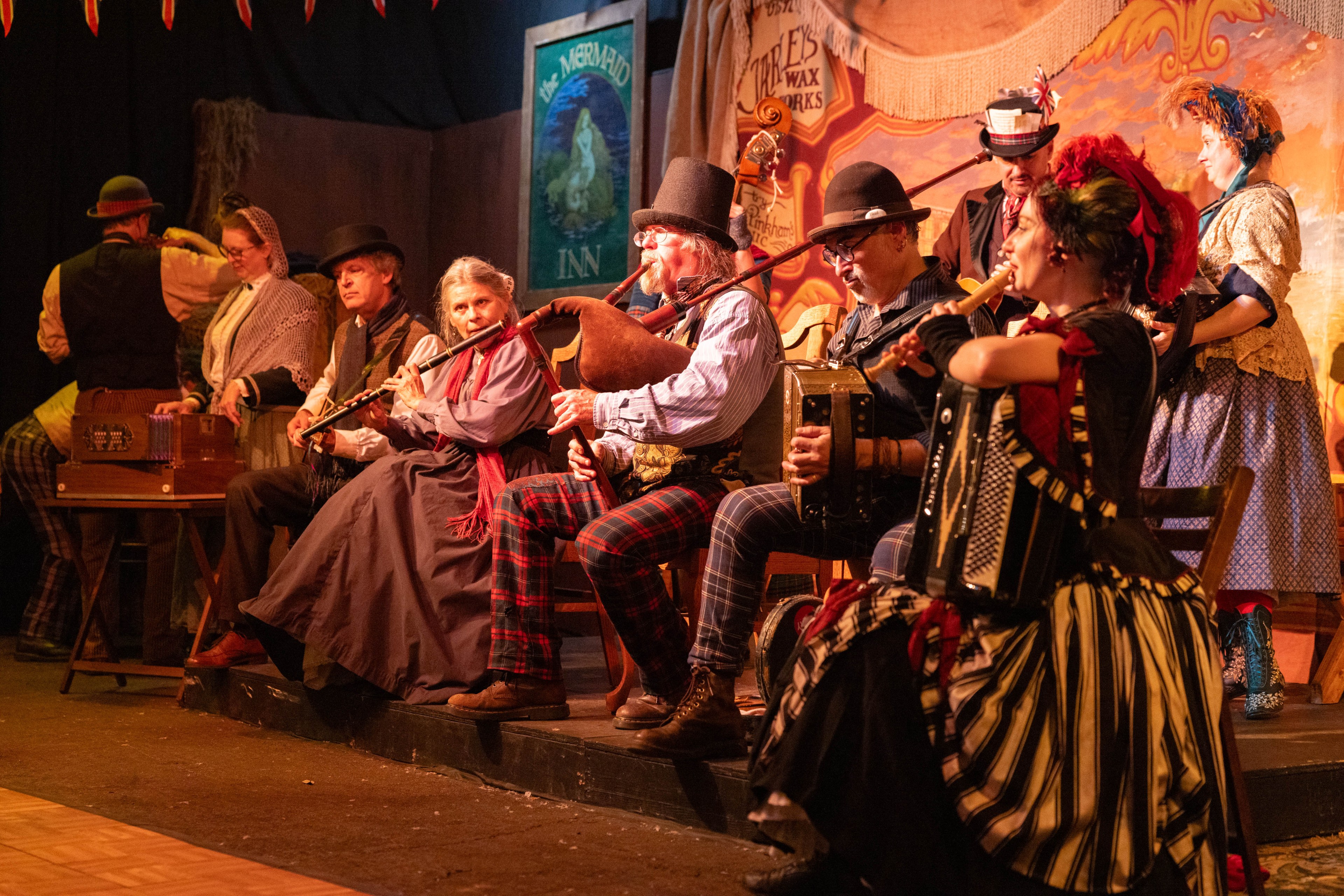 A group of people in vintage costumes play traditional instruments on stage, including a flute and accordion, under warm lighting, with a backdrop of vintage signs.