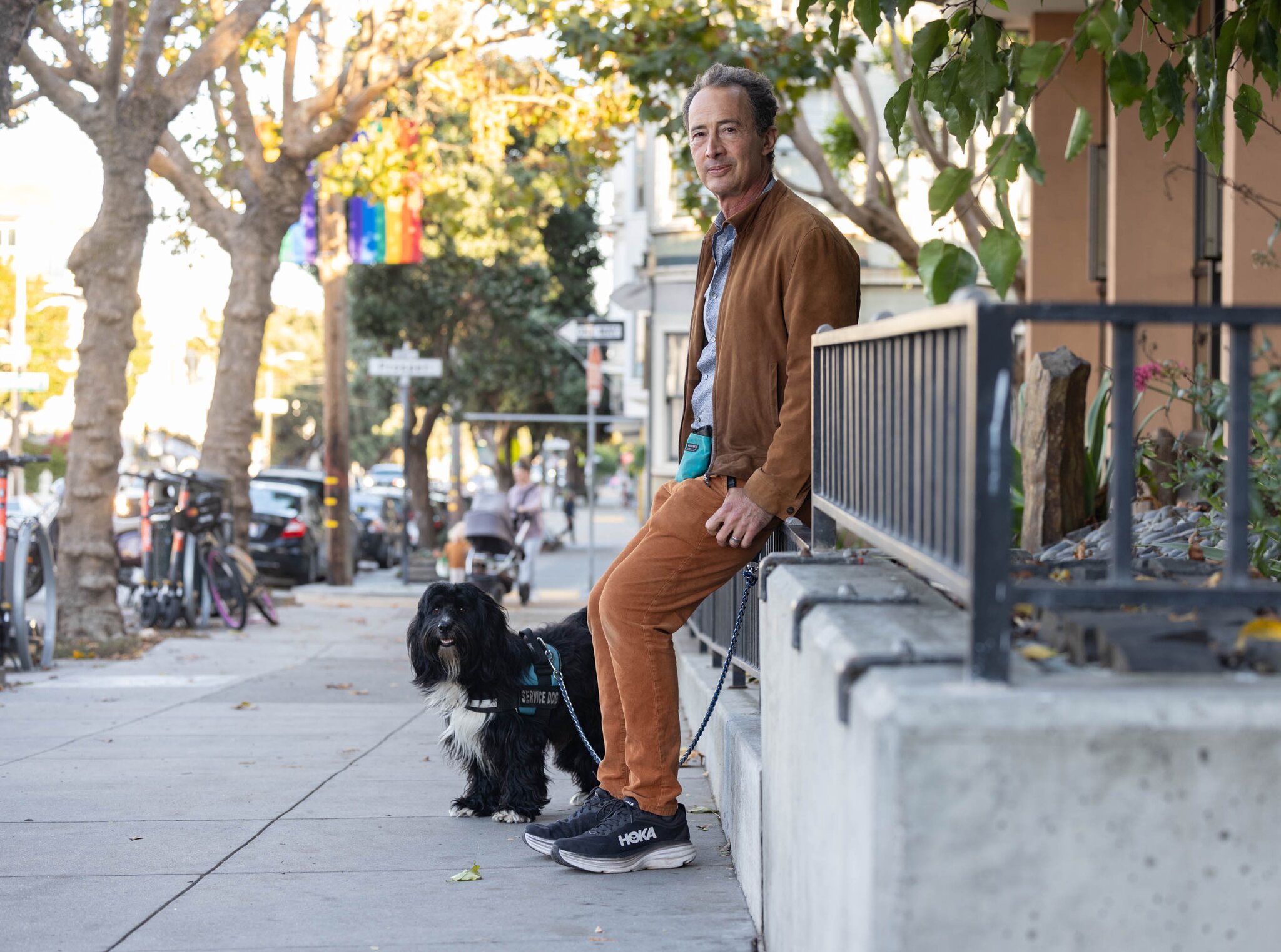 A small black dog lies on a sidewalk at the foot of its owner, who is wearing black sneakers and orange pants and shirt.