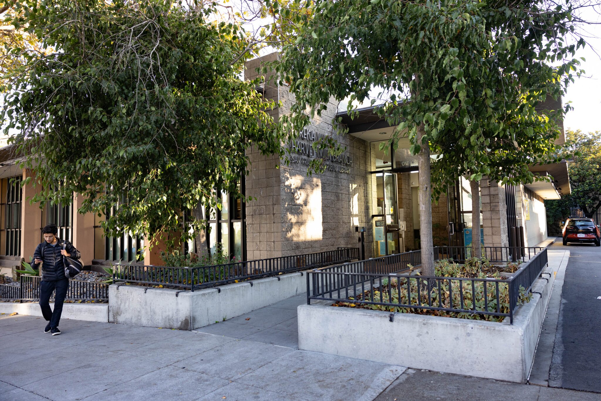 A pedestrian walks near a grey building's entrance, which has trees planted near it.