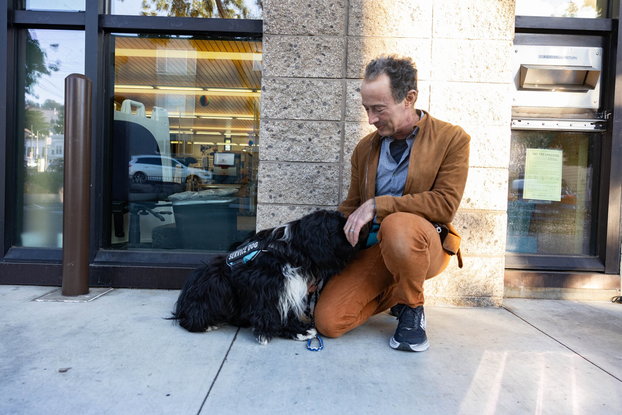 A small black dog lies on a sidewalk as it is petted by its owner, who is wearing black sneakers and orange pants.