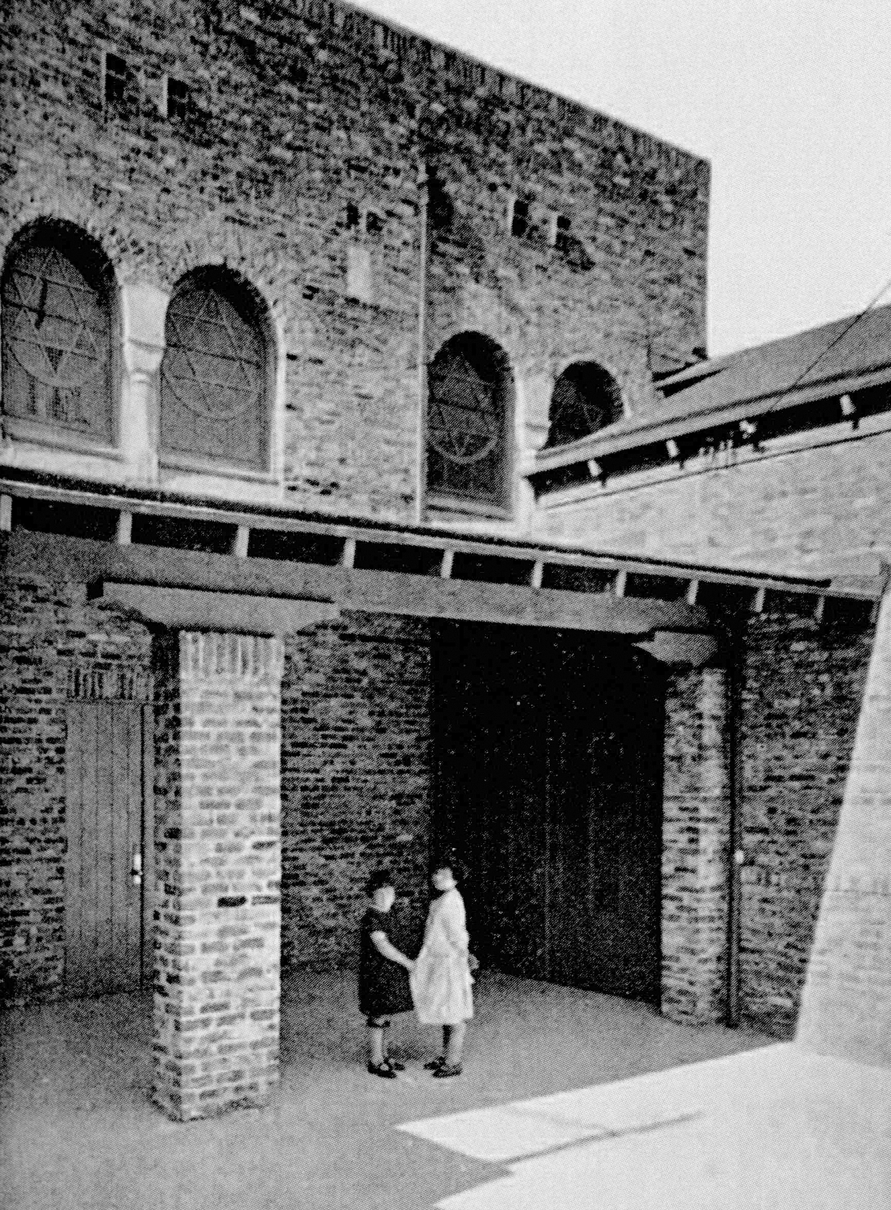 A black and white historical picture shows two people standing outside the Alper Memorial Building, the Jewish synagogue.
