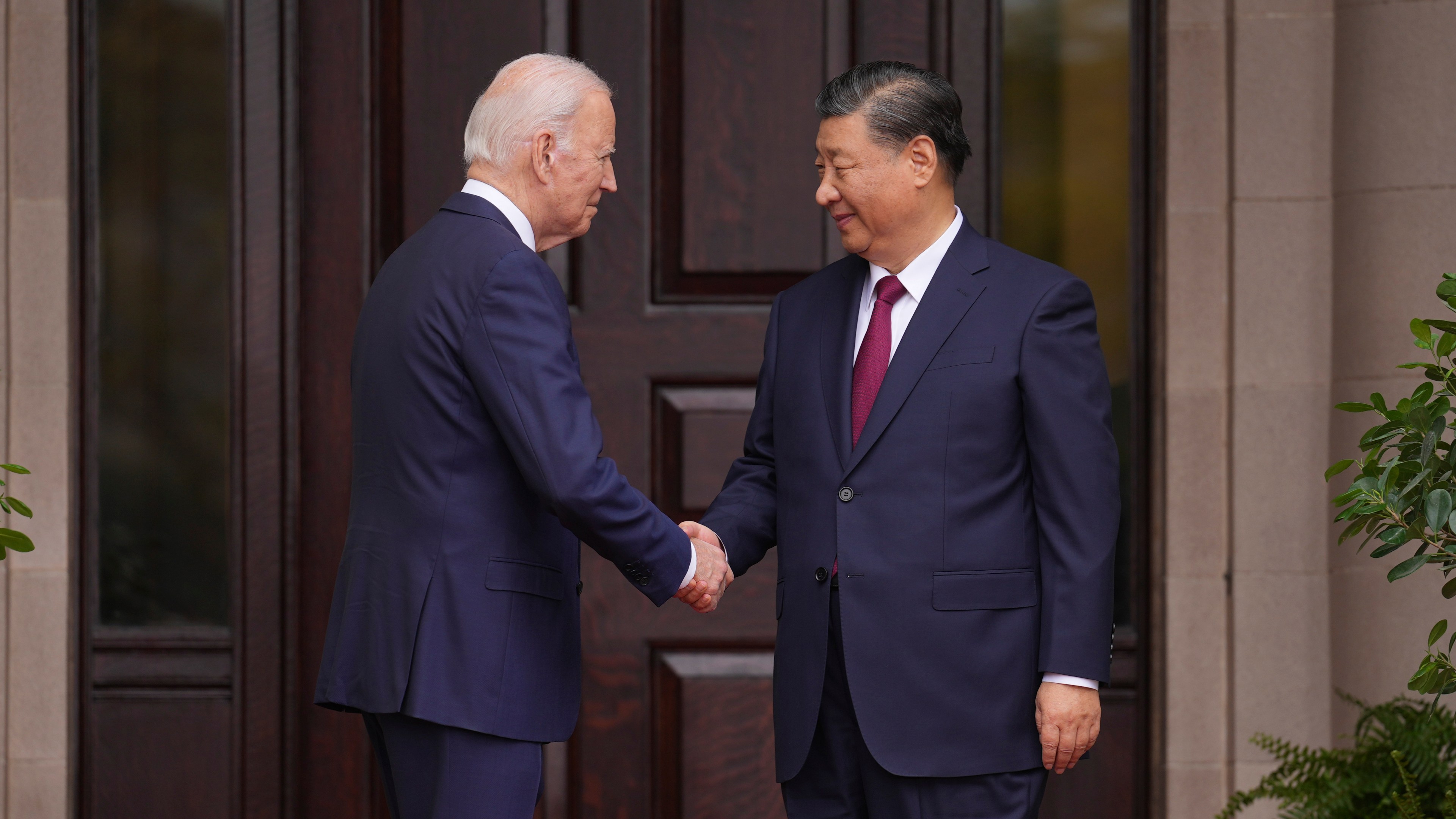 President Joe Biden shakes the hand of China's President President Xi Jinping next to a large wooden door.