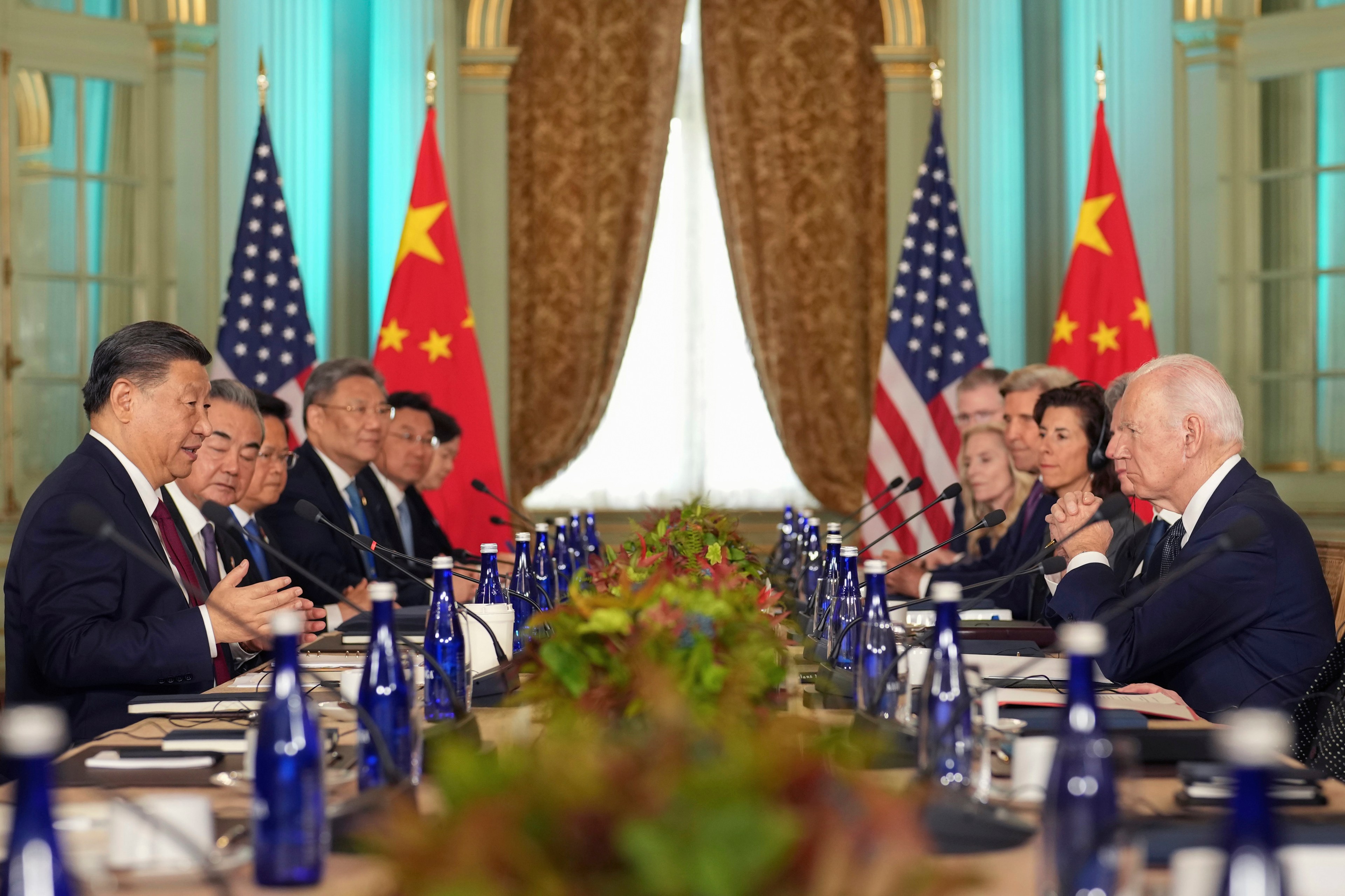 President Joe Biden listens as China's President President Xi Jinping speaks while seated at a long table with other delegates listening as well. Two Chinese and two American flags are at the back of the room in a formal dining room setting.