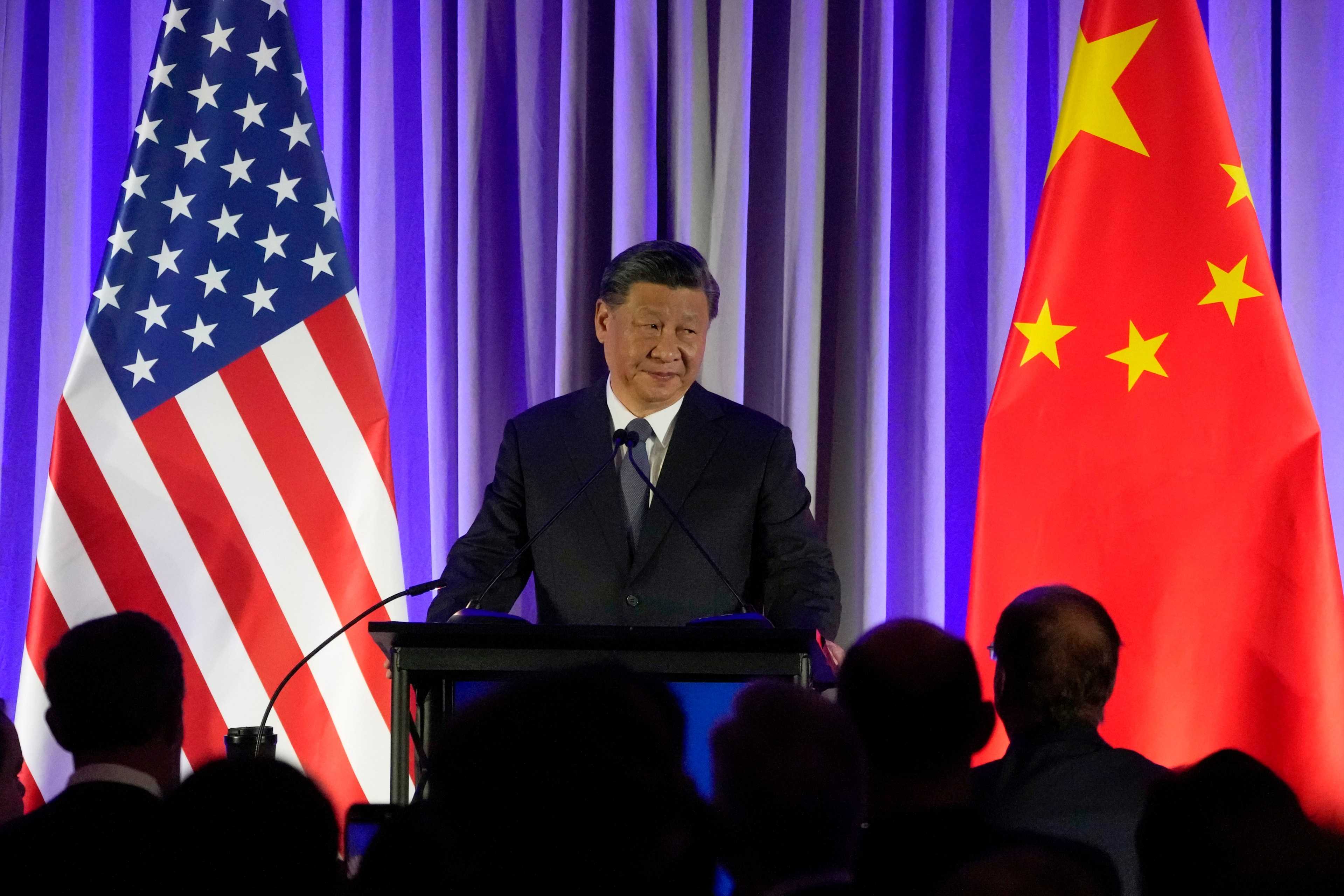 China's President Xi Jinping speaks to a crowd with a large American flag and Chinese flag on both sides of him.