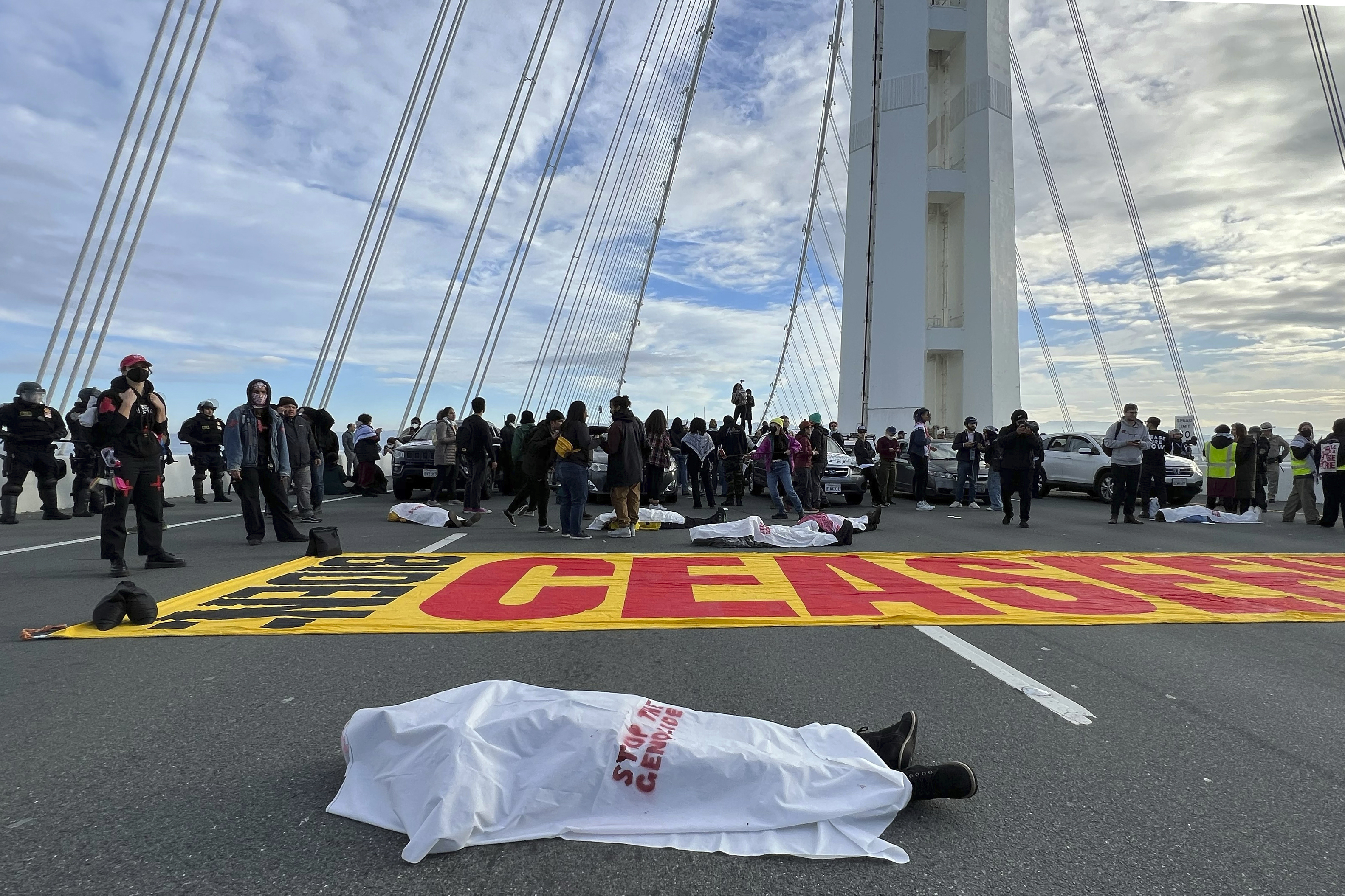 A person lays under a white sheet on a bridge with a large yellow banner that says CEASEFIRE with protesters blocking traffic.