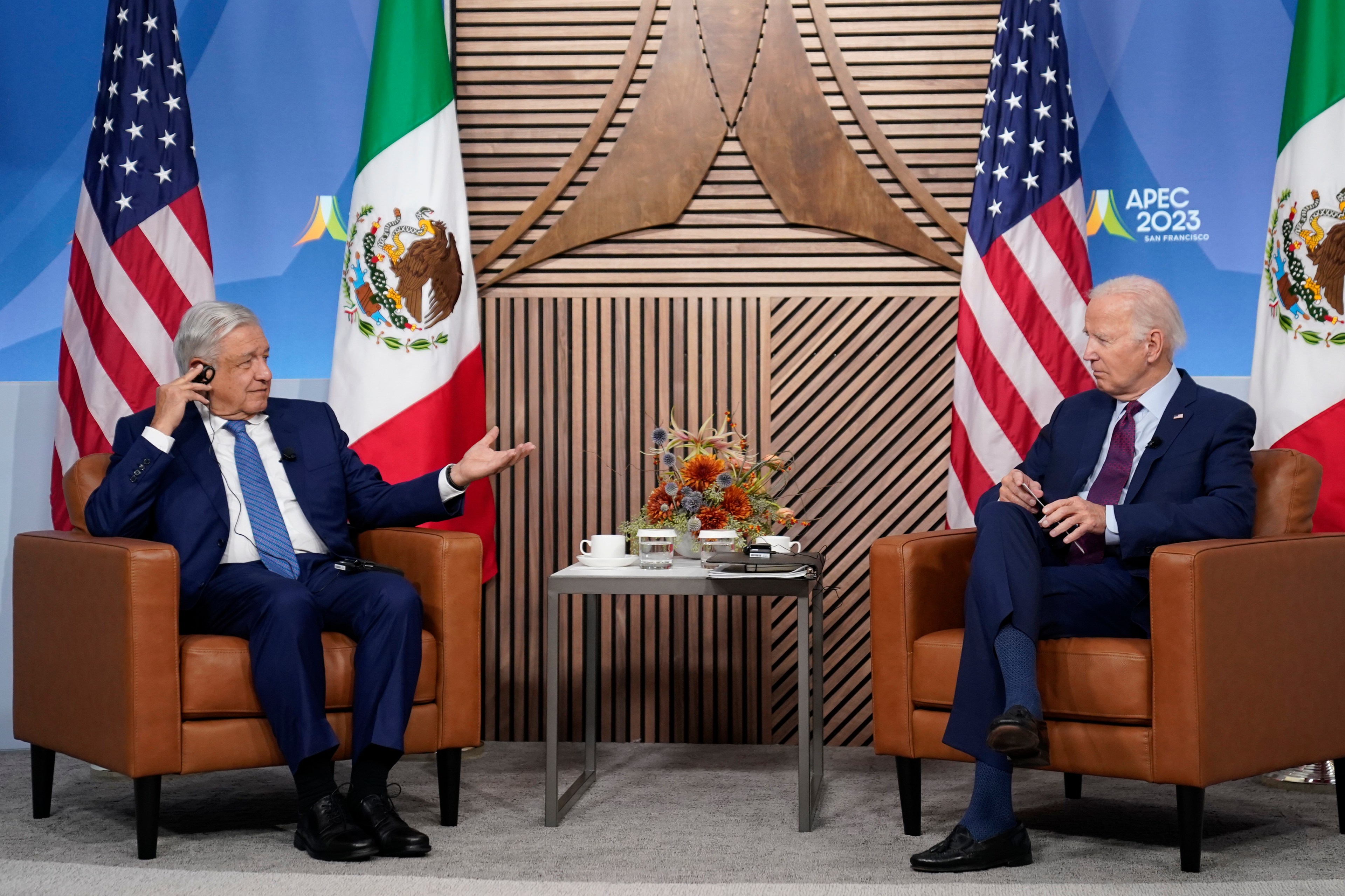 Two men in dark blue suits side in tan chairs with an end table between them. The man on the left sits in front of a Mexican flag. The man on the right, in front of a U.S. flag.