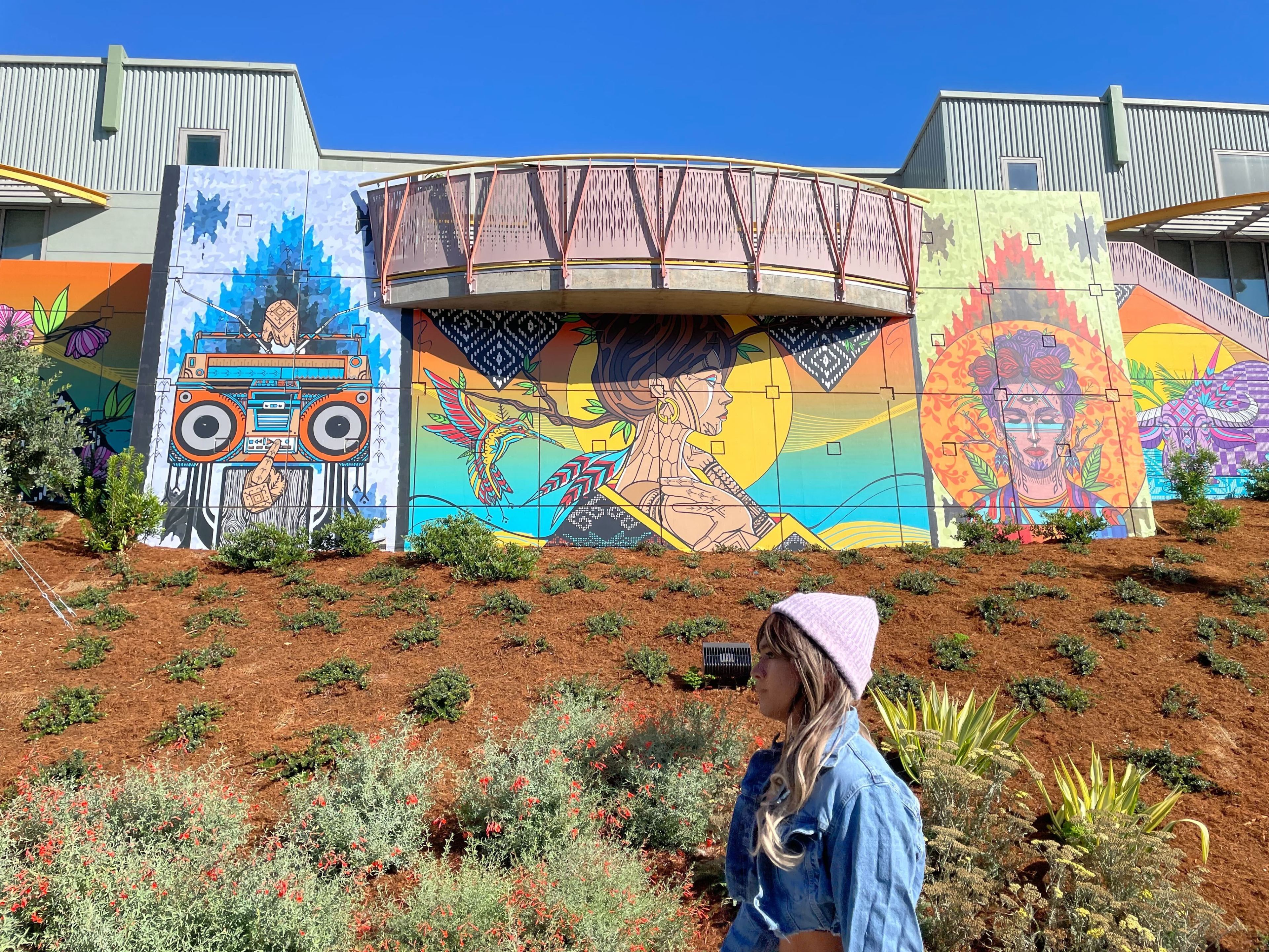 A woman in profile walks to the left in front of an embankment covered in wood chips with low-lying shrubs planted sparsely throughout it. Behind the embankment is a mural depicting, from left to right, a boombox, a woman's face in profile with stylized feathers next to it, and a woman with flowers in her hair and other designs facing the viewer.