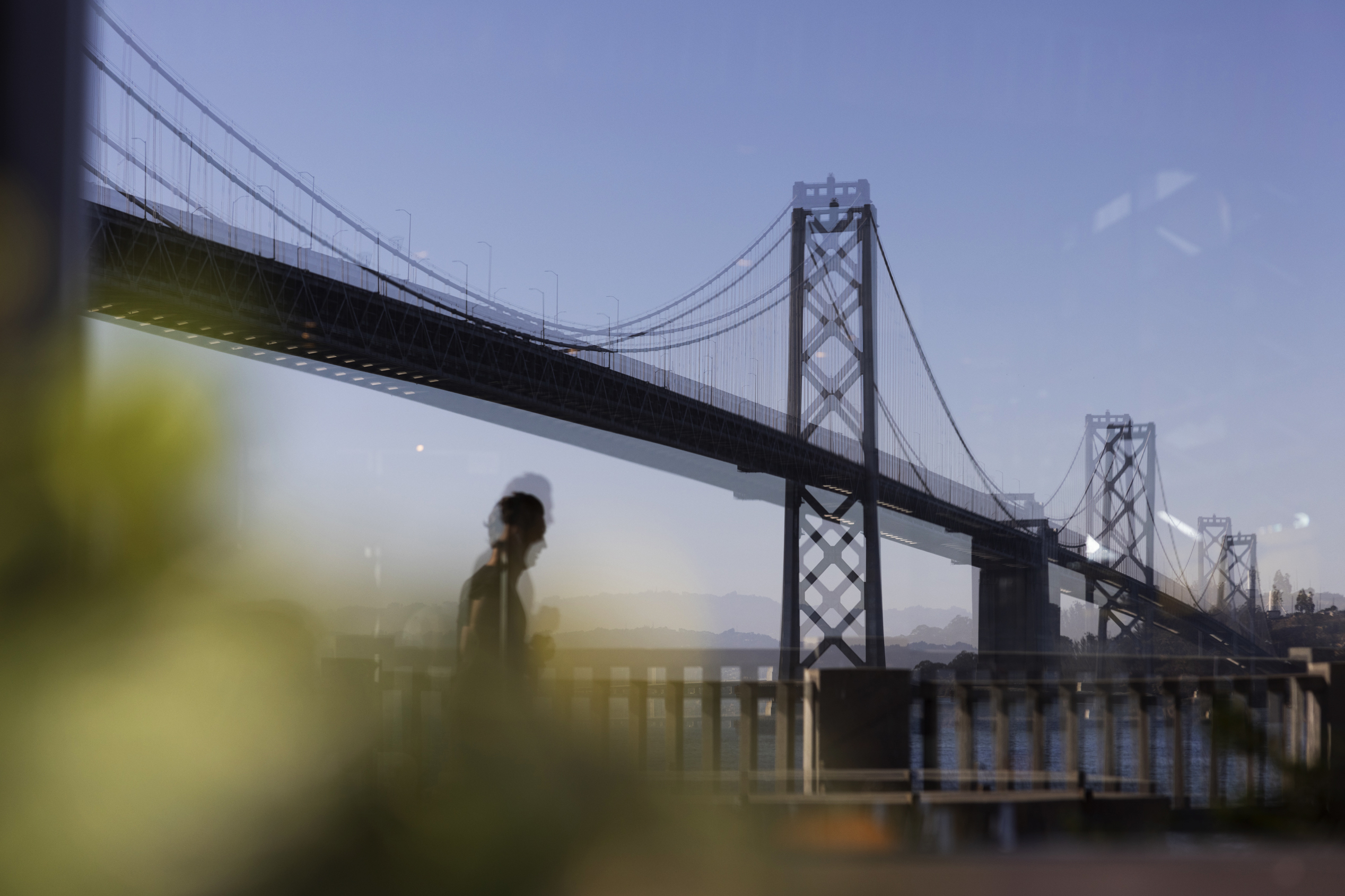 A silhouetted pedestrian walks by The San Francisco-Oakland Bay Bridge which is reflected in a window on a sunny afternoon.