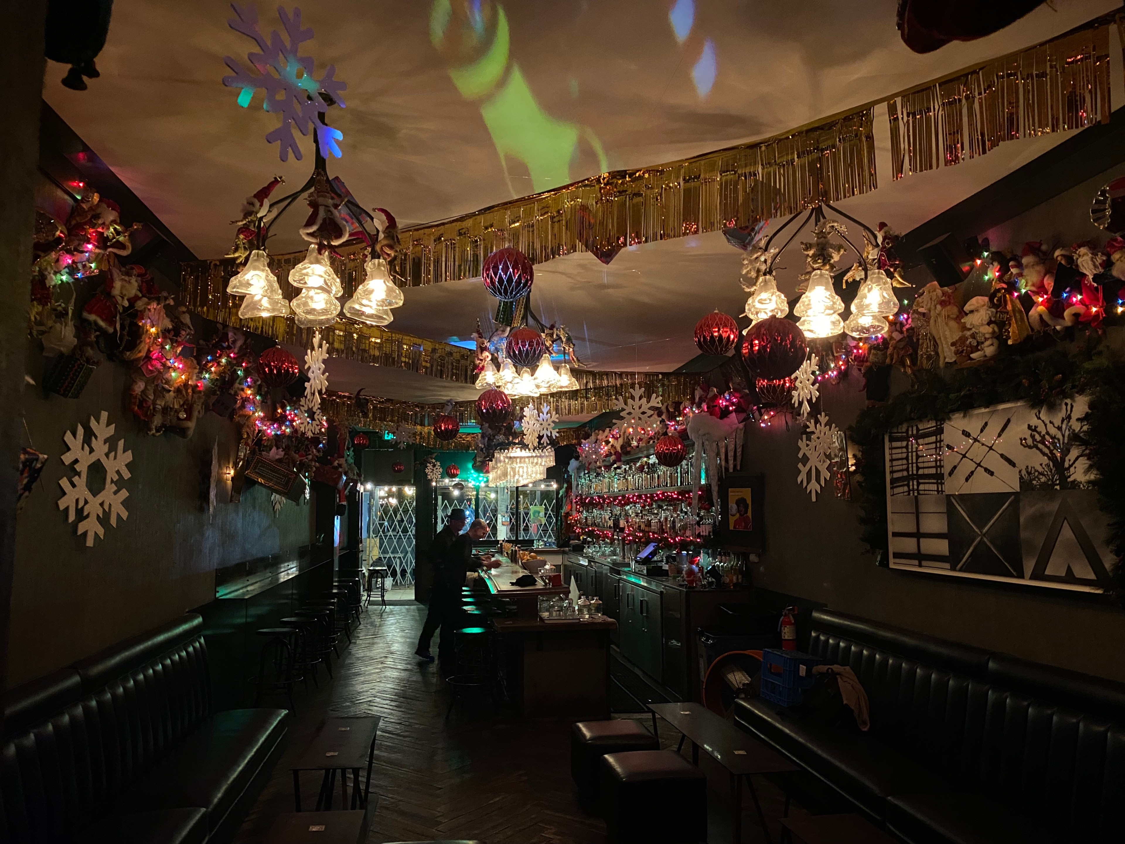 A dark bar with chandeliers is decorated with tinsel, glittering garlands, snowflake cut-outs and hundreds of vintage Santa dolls.  
