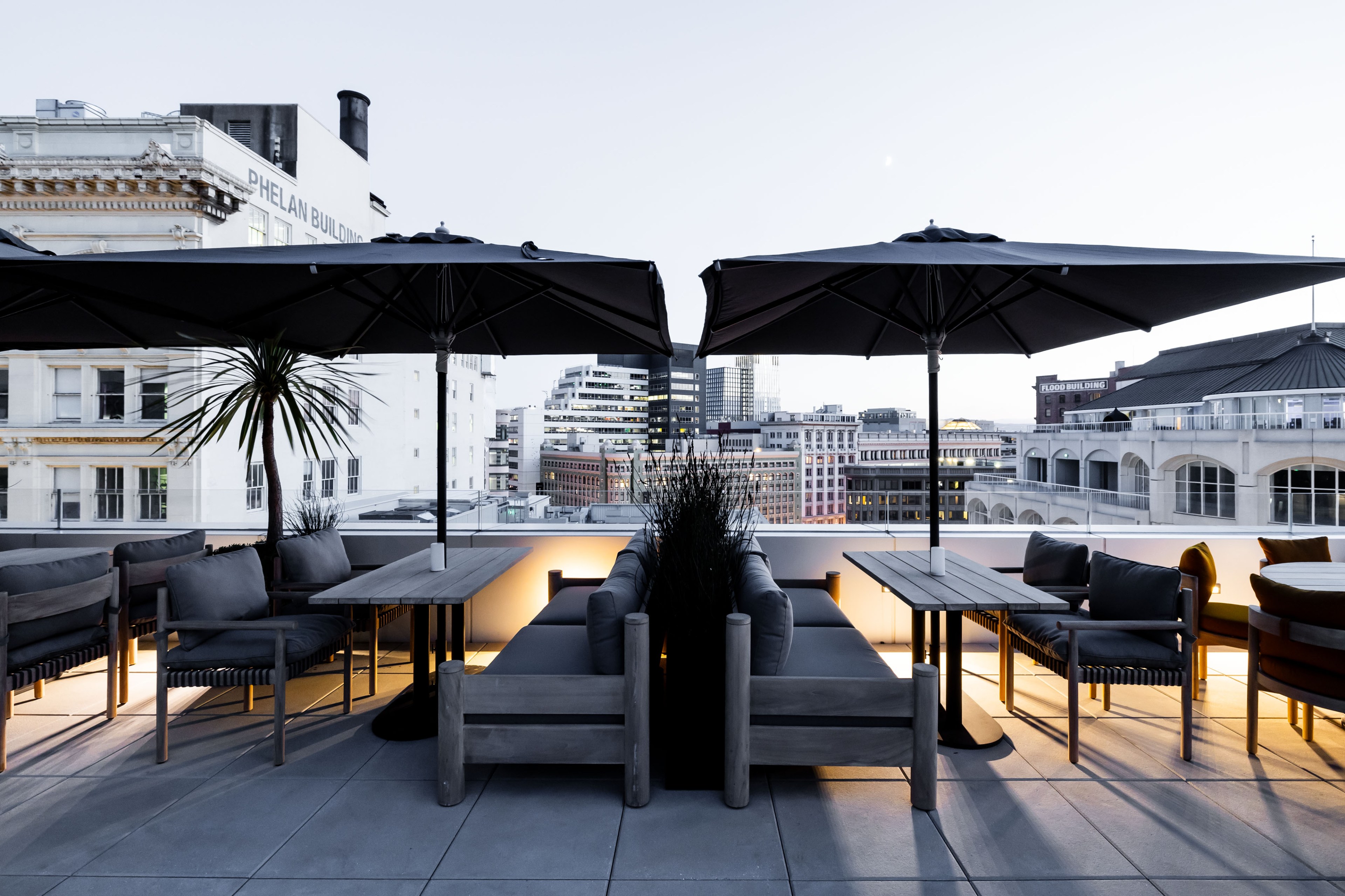 Tables with umbrellas stand on a rooftop patio with the San Francisco skyline in the background.