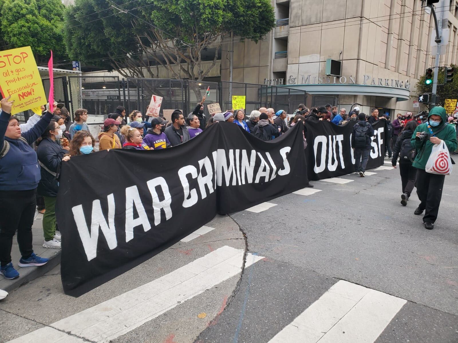A row of protesters holds a black banner with words in white block lettering with the words &quot;War criminals out of SF&quot; in front of tall black security fencing blocking off a street intersection.