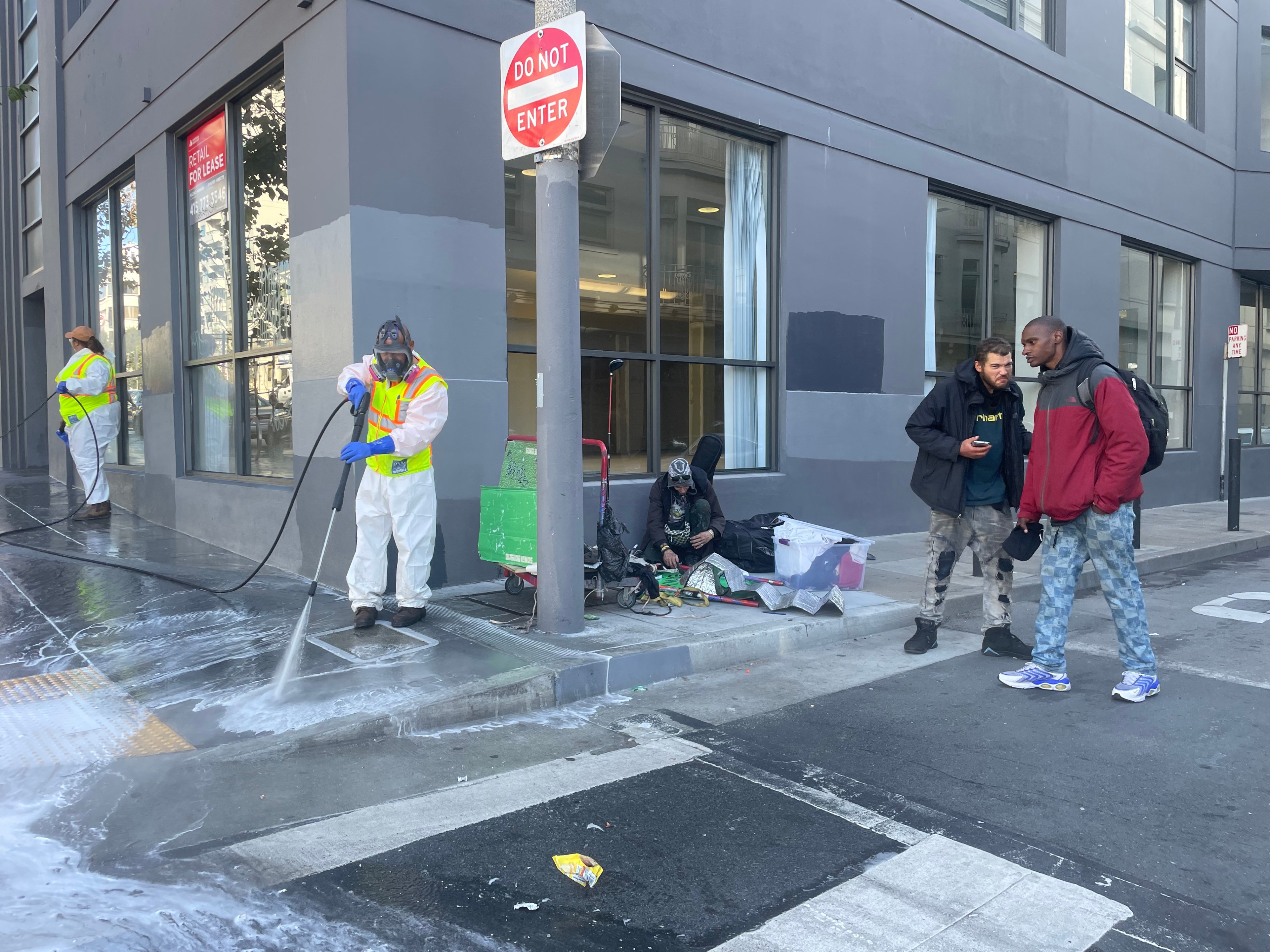 A city worker washes the sidewalk while unhoused people stand to the side.