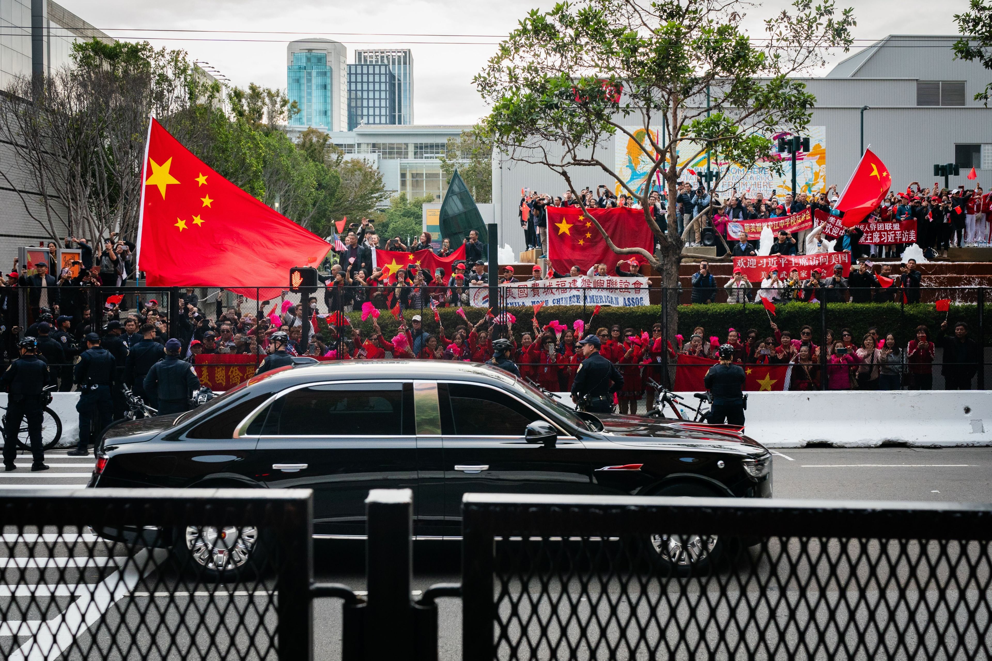 Chinese supporters wave flags behind a metal barricade on a city street as a black sedan passes by.