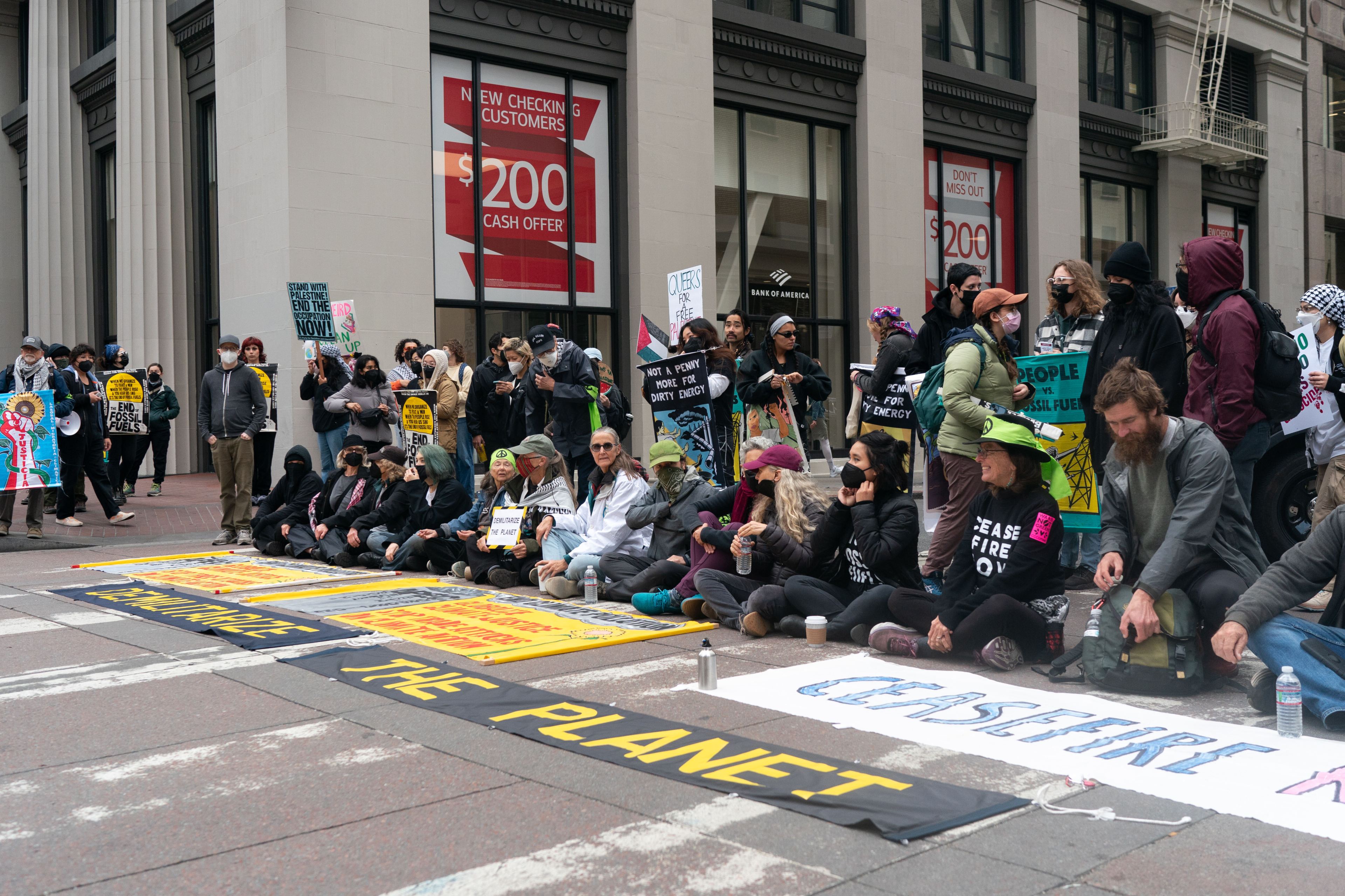 A long line of protesters side on the street blocking traffic with a &quot;Demilitarize the planet&quot;banner on the ground.