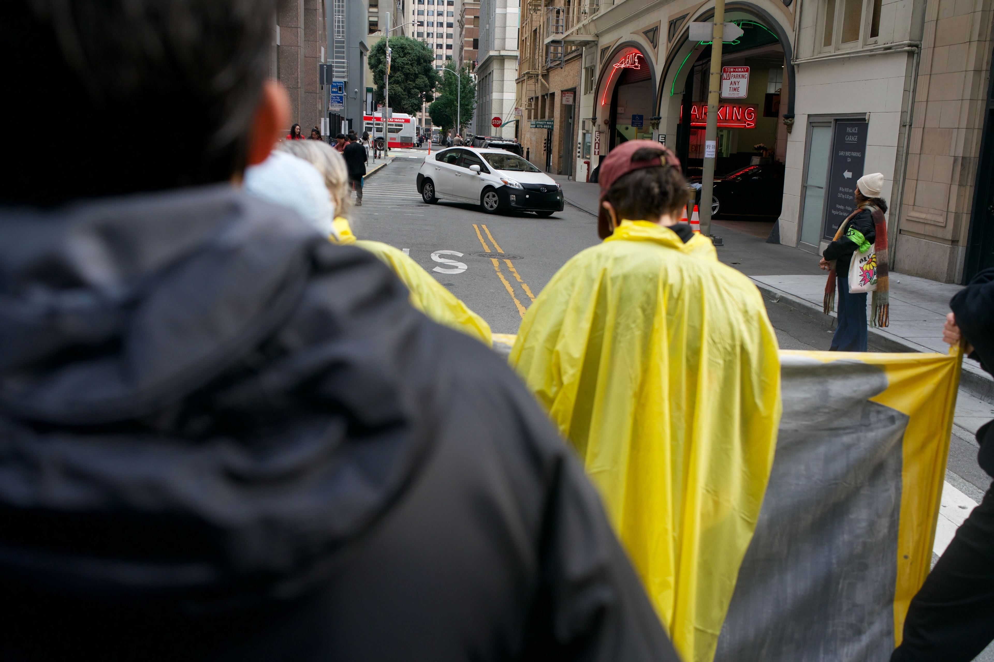 a white car reverses down a blocked street as poncho-clad protesters look on.