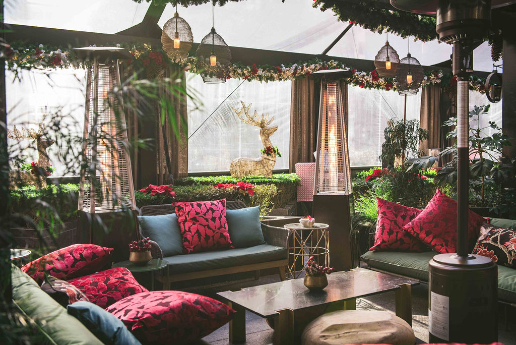 A tented lounge with a clear ceiling features dangling lights and green couches with bright red pillows for the holidays. 