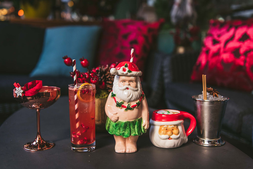 A lineup of holiday-themed cocktails, including drinks in brass martini glass, a Santa-shaped mug, and a cup with Santa wearing a hula skirt.
