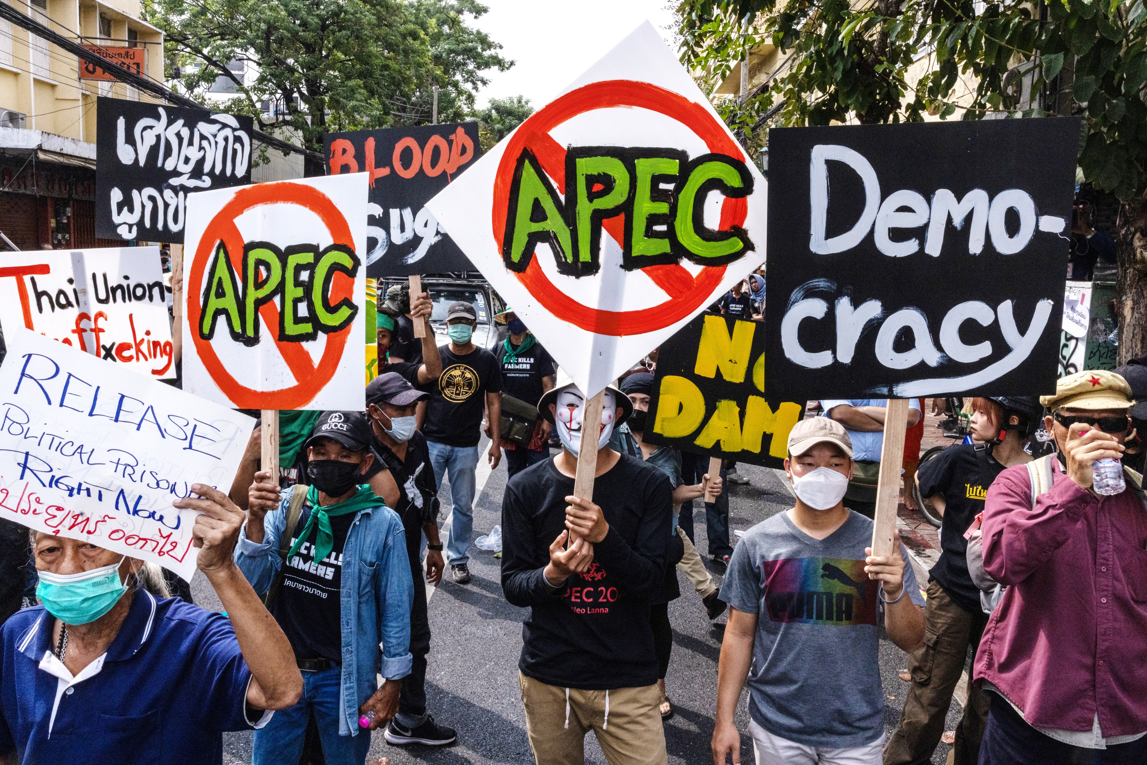 Dozens of people wearing COVID face mask stand closely together in the middle of a street in Bangkok, Thailand, during an Anti-APEC protest. They are holding signs that read "Democracy" "Release Political Prisoners Right Now" and the acronym "APEC" with a big red circle and strike through the words.