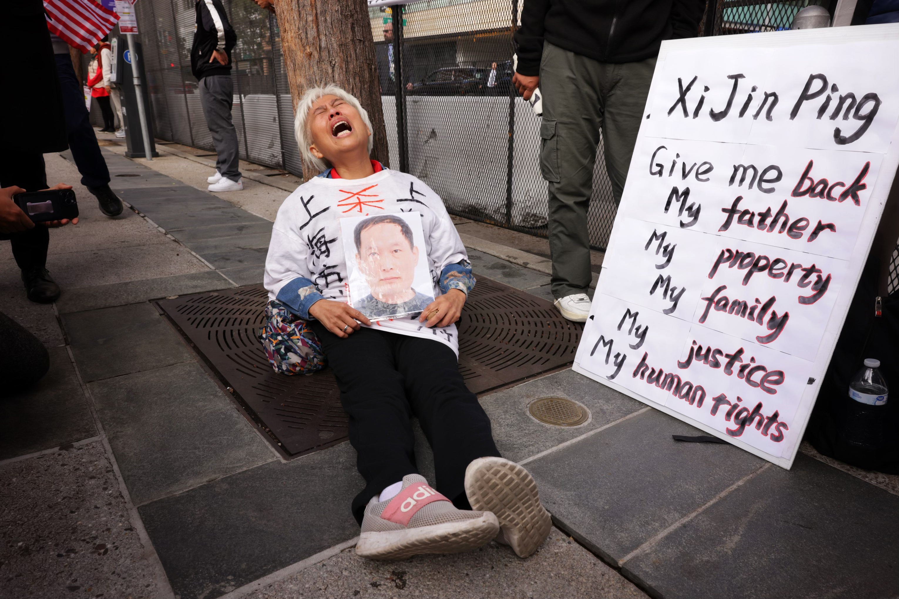 A woman screams while sitting on the ground with a large sign next to her that reads "Xi Jin Ping, Give me back My father. My property, My family, My justive, My human rights"