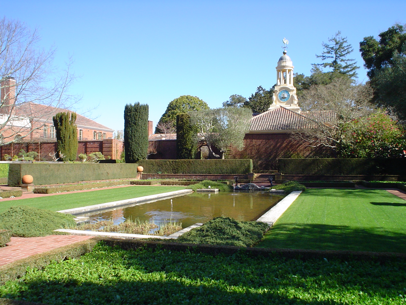 A large outdoor garden area with a large sunken pool and a large brick estate in the background on a sunny day.