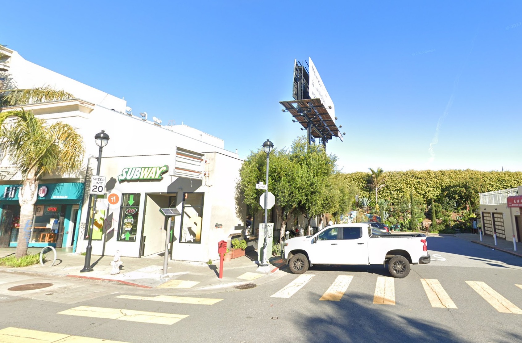 A sunny street intersection under blue skies with a white pickup truck parked outside one of several businesses.