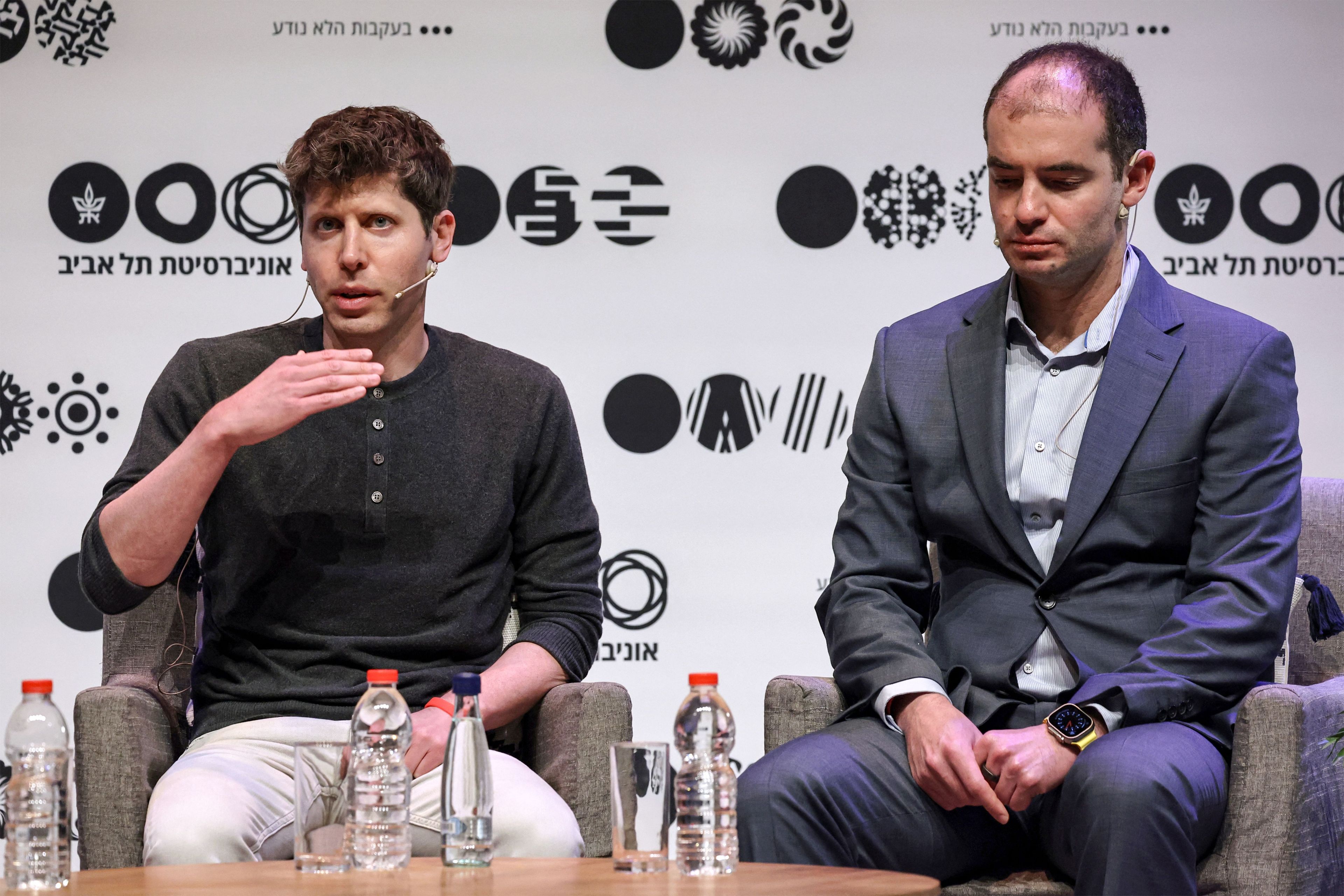 Sam Altman wears a gray shirt while talking at Tel Aviv University. His right arm is raised up. Ilya Sutskever, to Altman's left, is looking down.