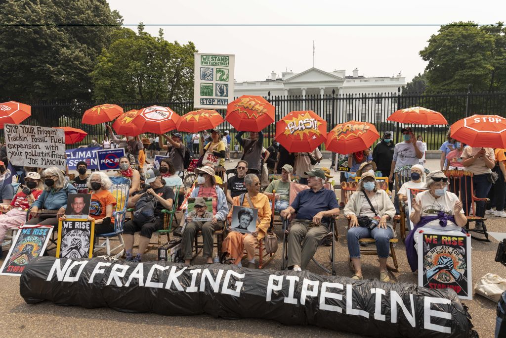 A group of more than two dozen people sit in front of the White House with a black cylindrical sign reading &quot;no fracking pipeline.&quot; The protesters are also holding red umbrellas and signs protesting the Mountain Valley Pipeline, which is used for transporting fossil fuels. 