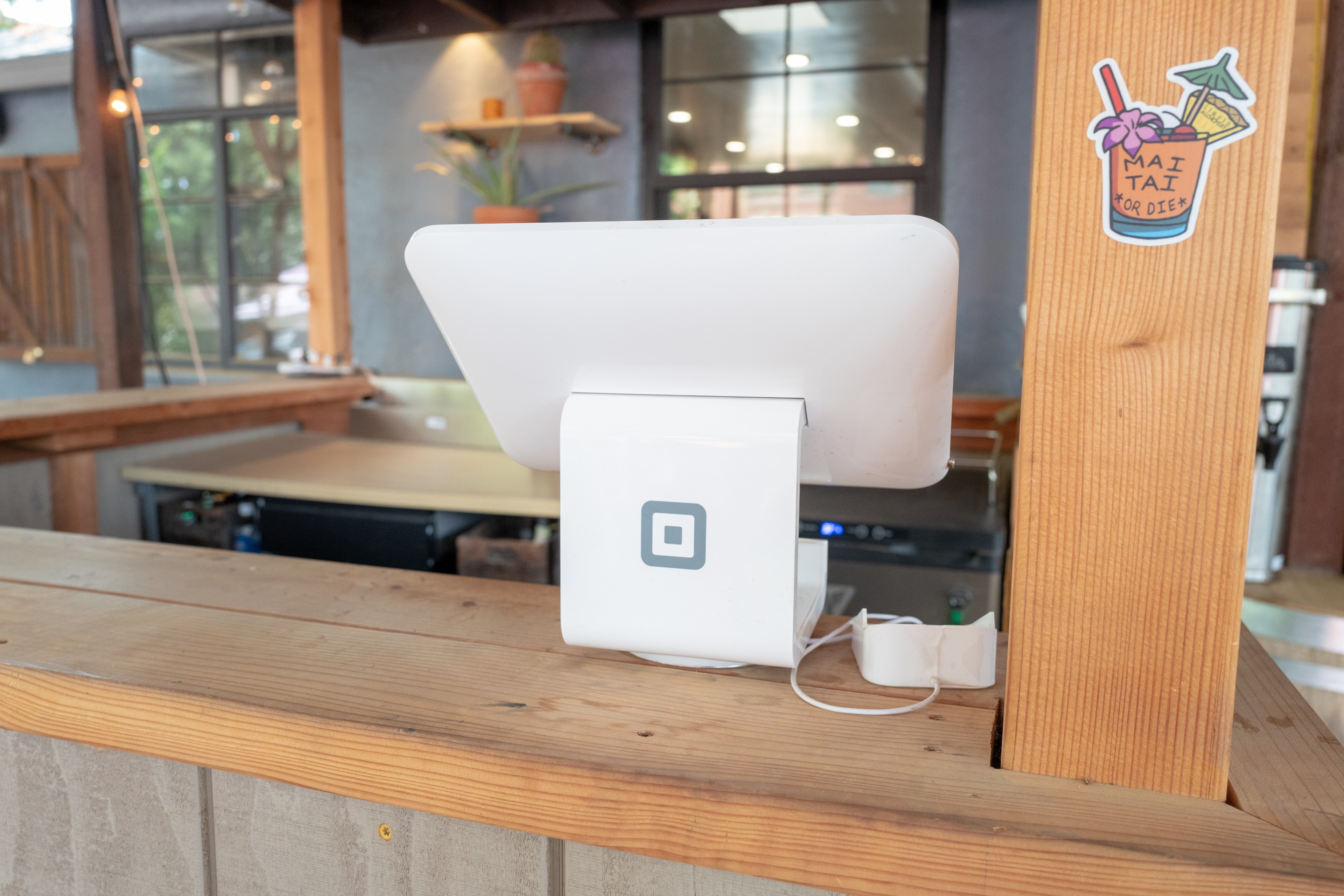A white Square payment terminal placed on a wood counter. A sticker of a tiki drink with the words "mai tai" printed on it is placed on a column next to the counter.
