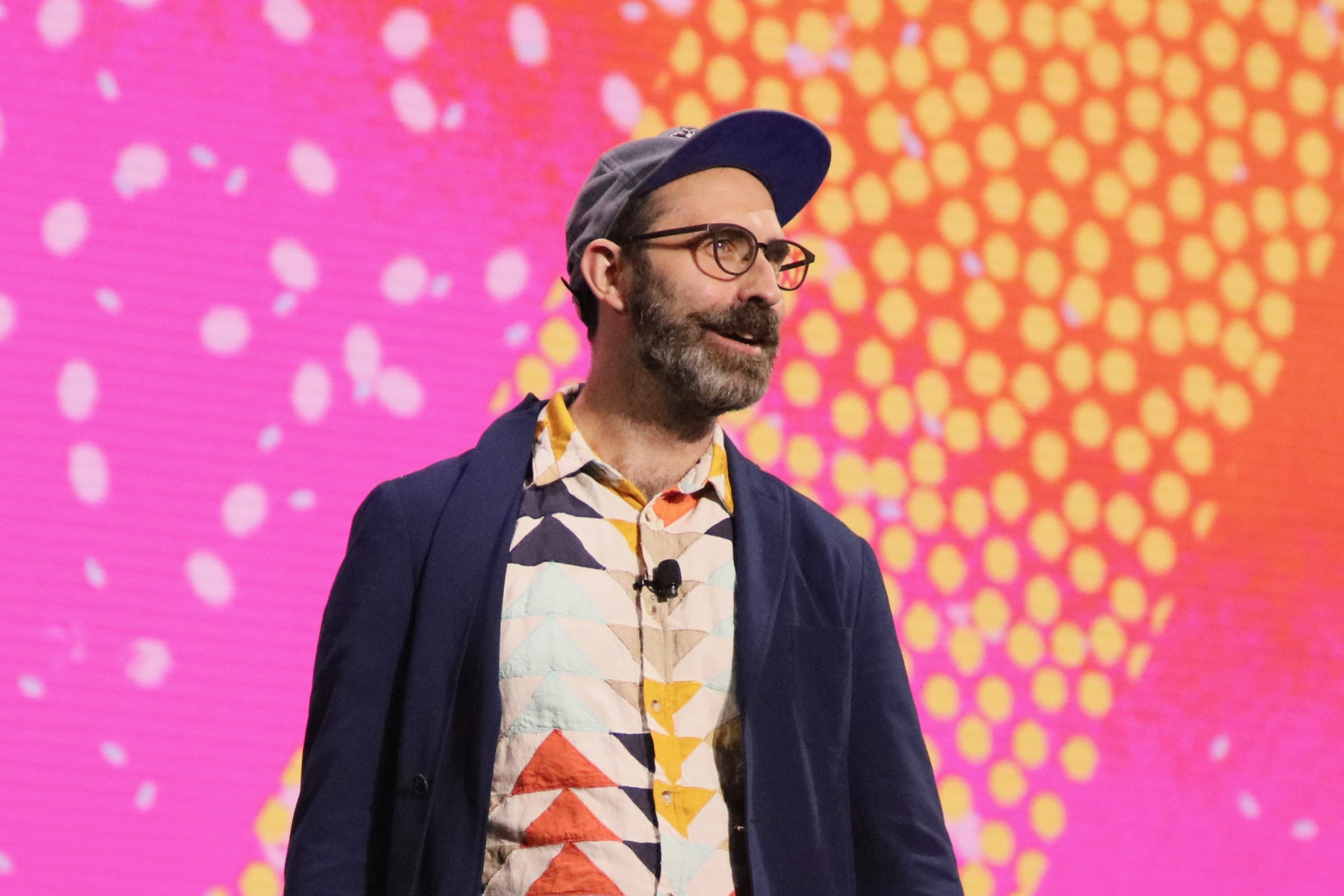 A man with a beard, glasses and ball cap looks into the distance with a pink and orange artful backdrop behind him