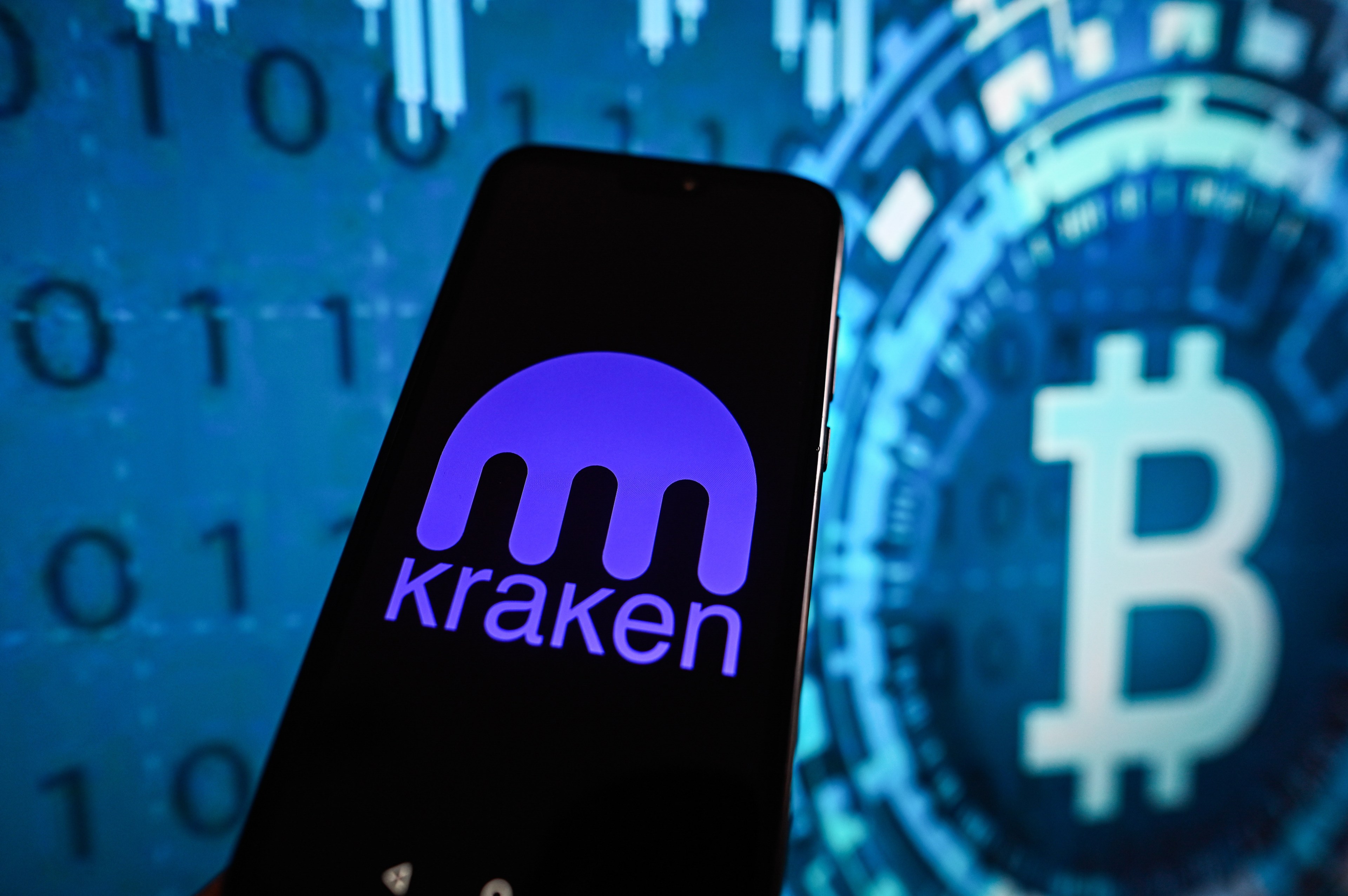 A Kraken logo displayed on a smartphone with a Bitcoin logo in the background.