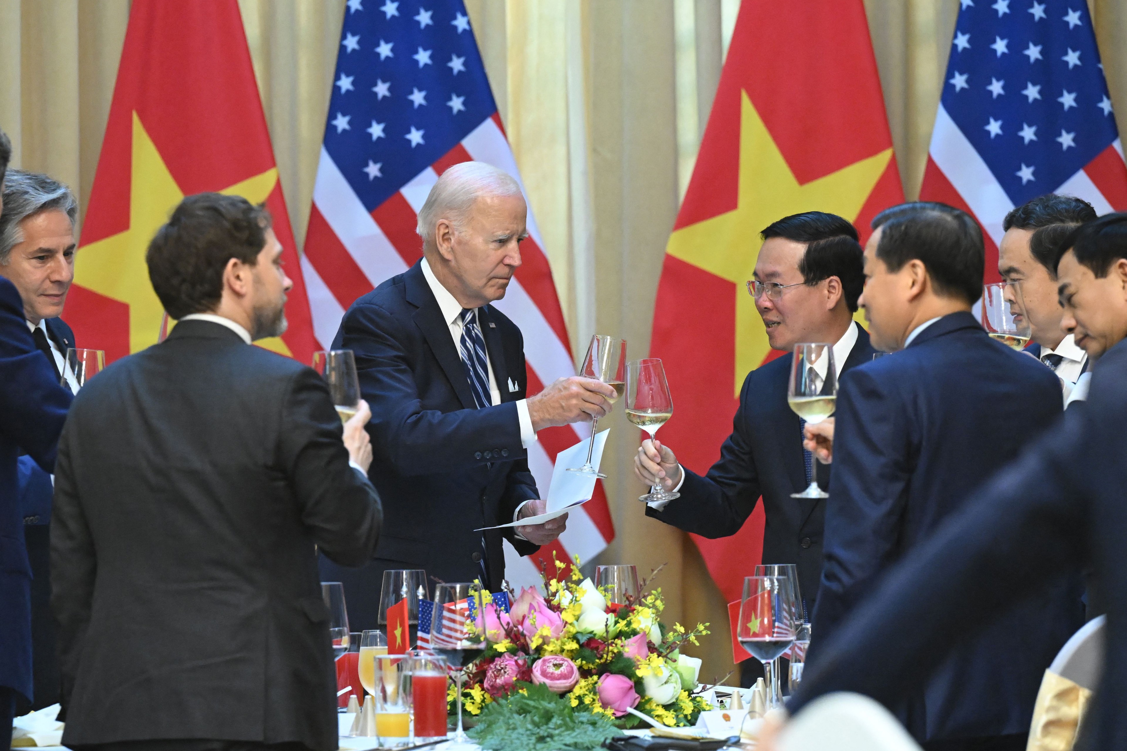 Surrounded by standing men in suits and 2 American flags and 2 Vietnam flag, US President Joe Biden stands in the center of the picture holding a glass of wine as he prepares to cheers with Vietnamese President Vo Van Thuong during a formal dinner setting.
