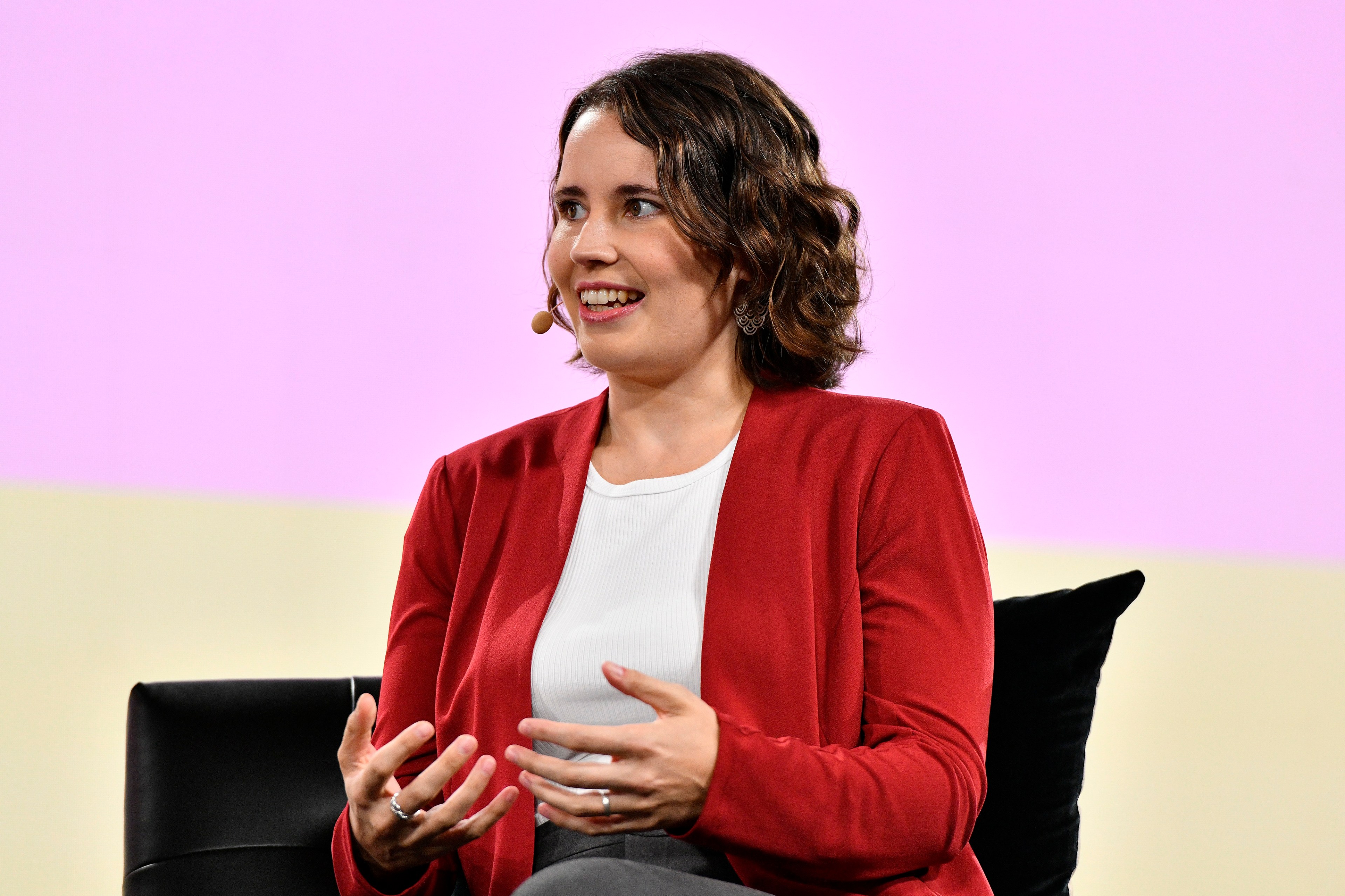 Helen Toner of Georgetown's CSET wears a red cardigan and a white blouse speaking at Vox Media's 2023 Code Conference. Behind her is a pink and yellow background.