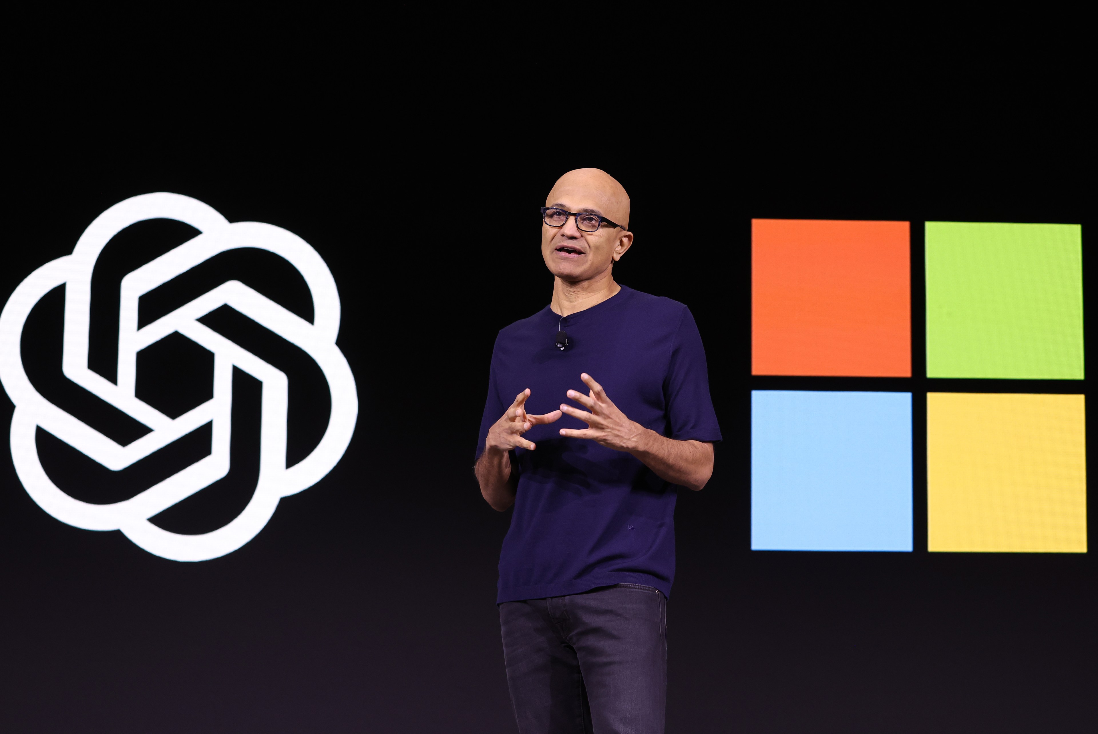 Microsoft CEO Satya Nadella speaks while on stage betwen the OpenAi logo and the Microsoft logo