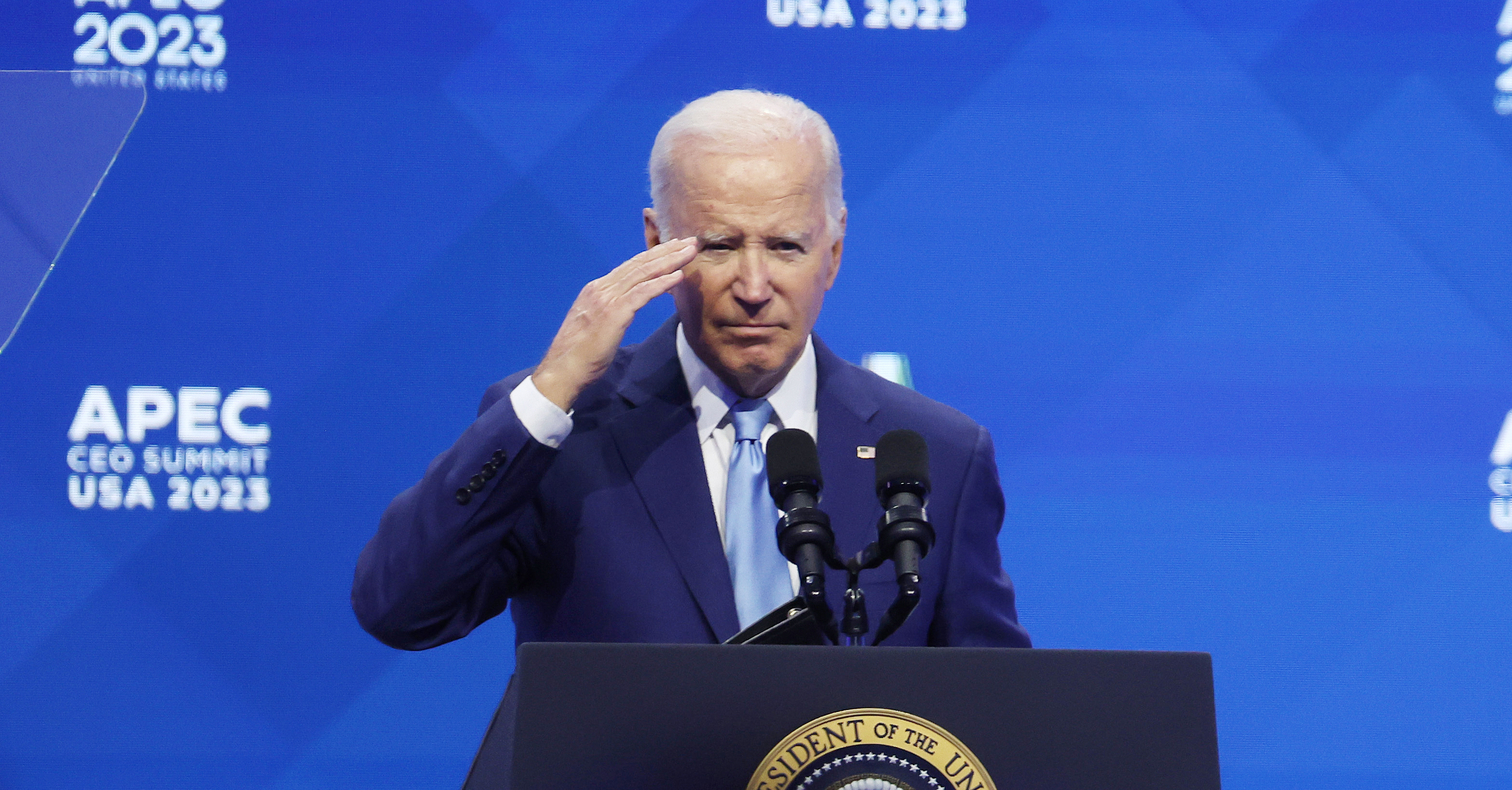 U.S. President Joe Biden stands behind a lectern saluting with a blue backdrop in the background that reads: APEC CEO SUMMIT USA 2023