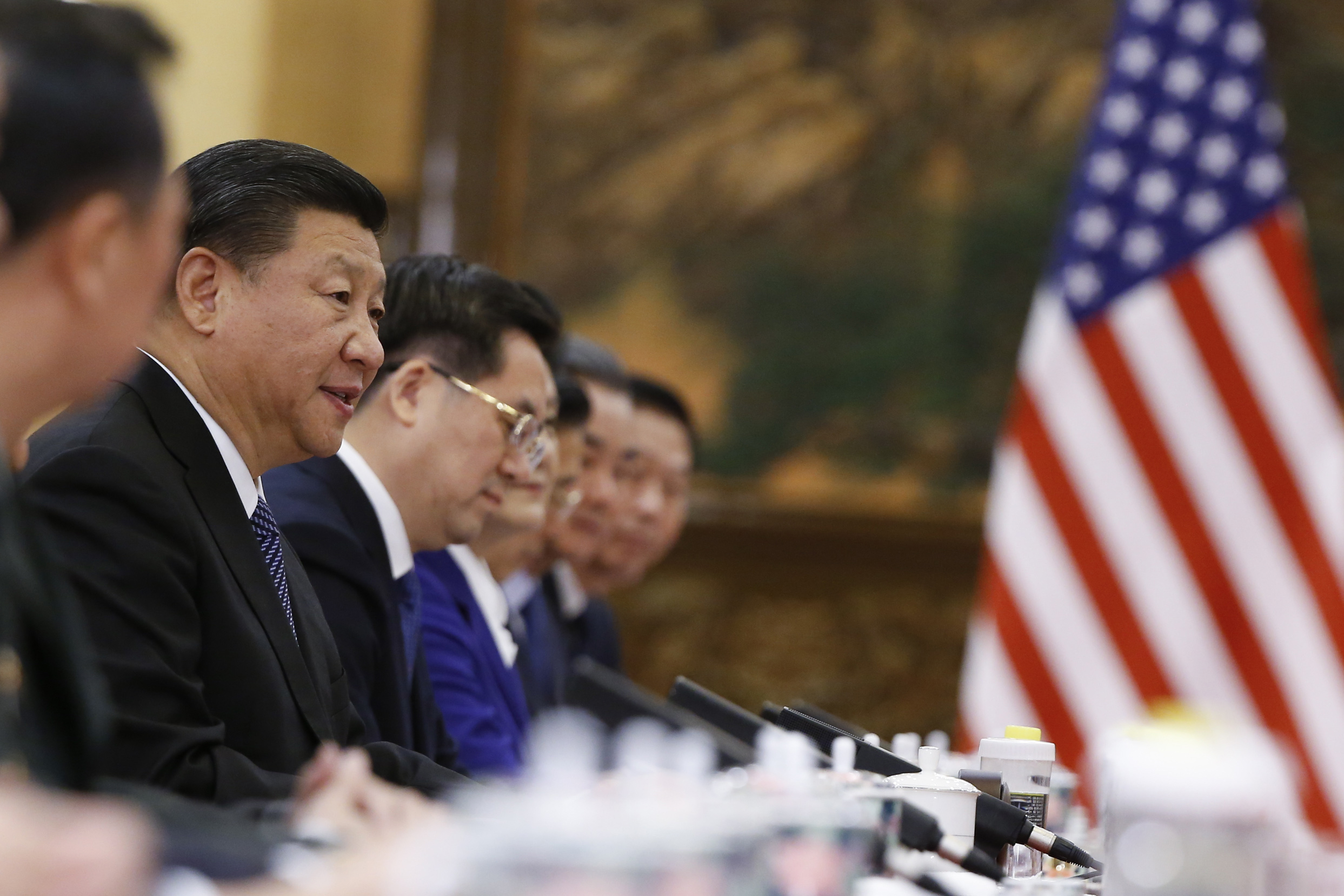 China's President Xi Jinping sits at a long table with other people with the American flag in the background