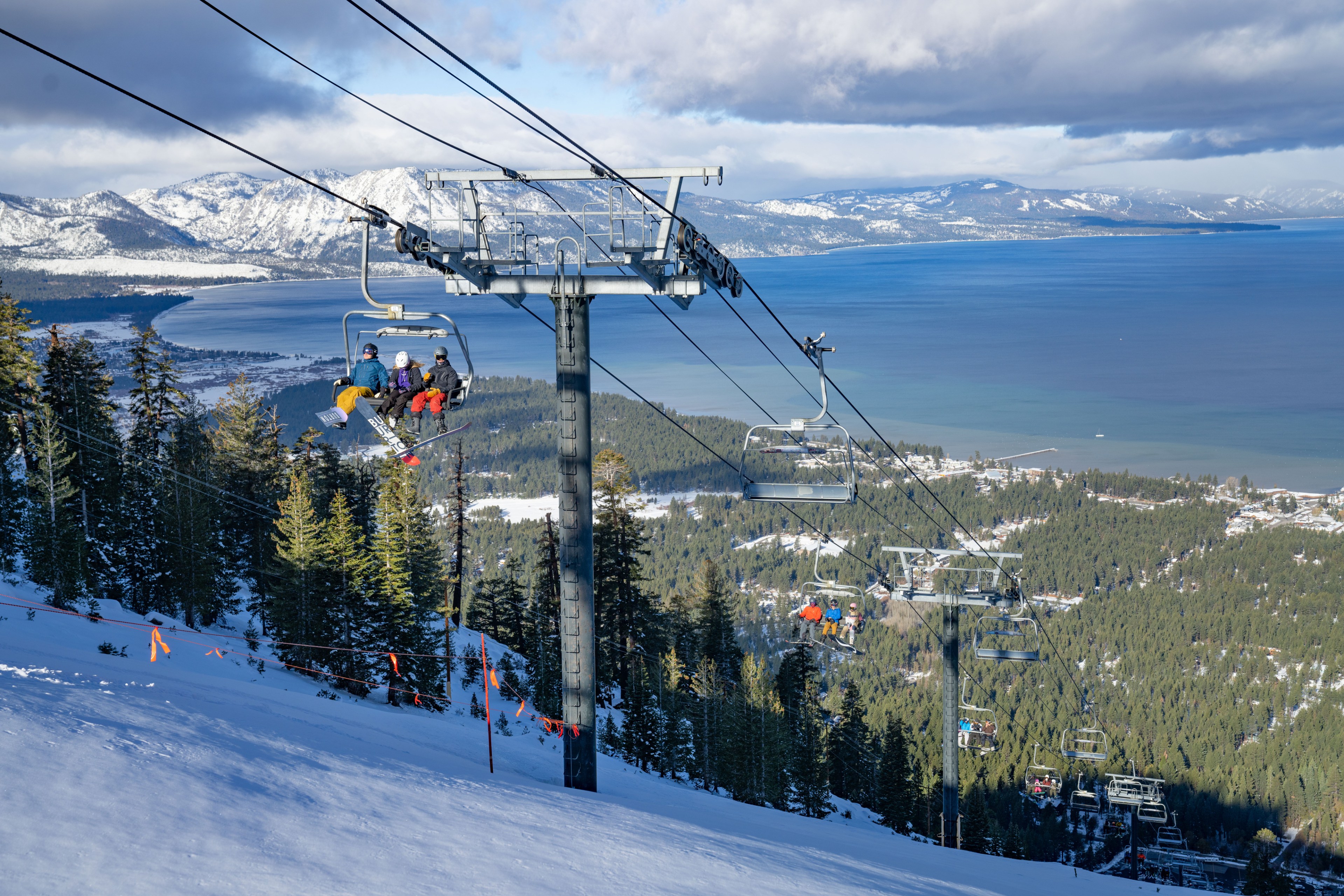 Skiiers ride a lift overlooking a snowy mountain and a lake in the distance.