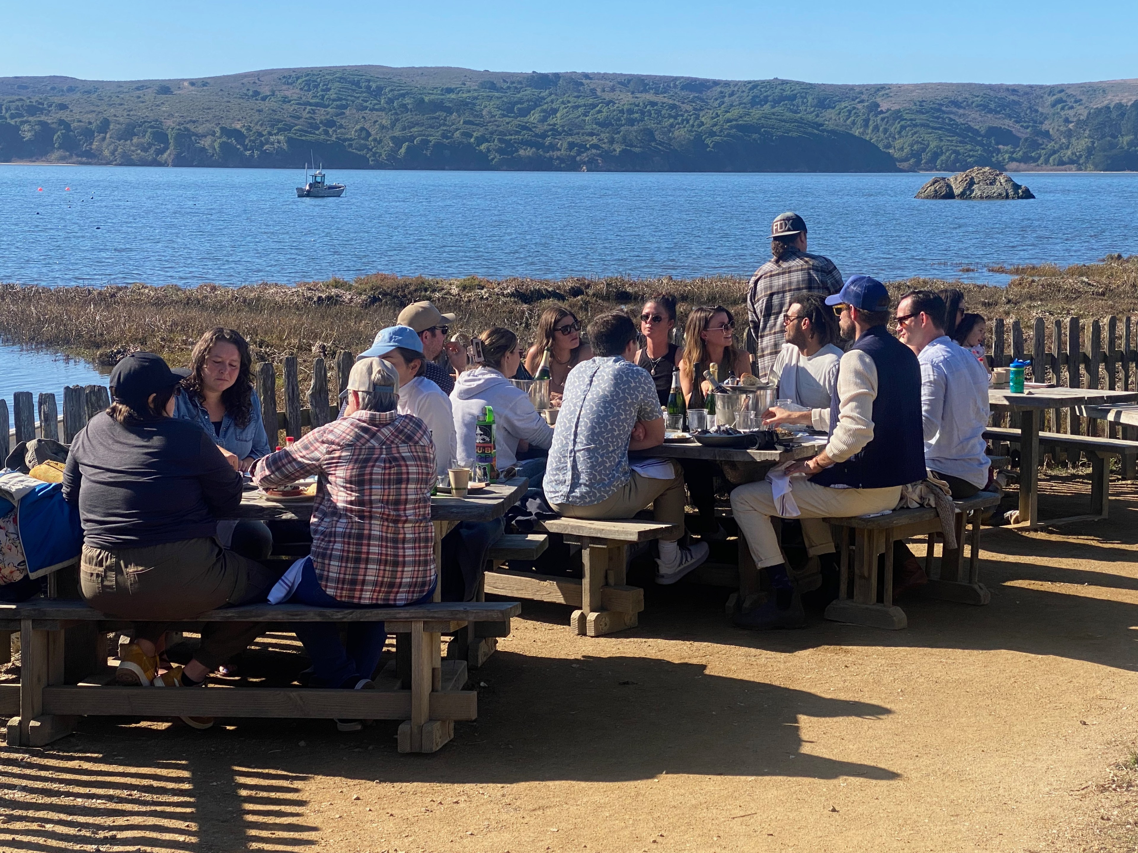 People gather eating oysters at picnic tables next to the sea.