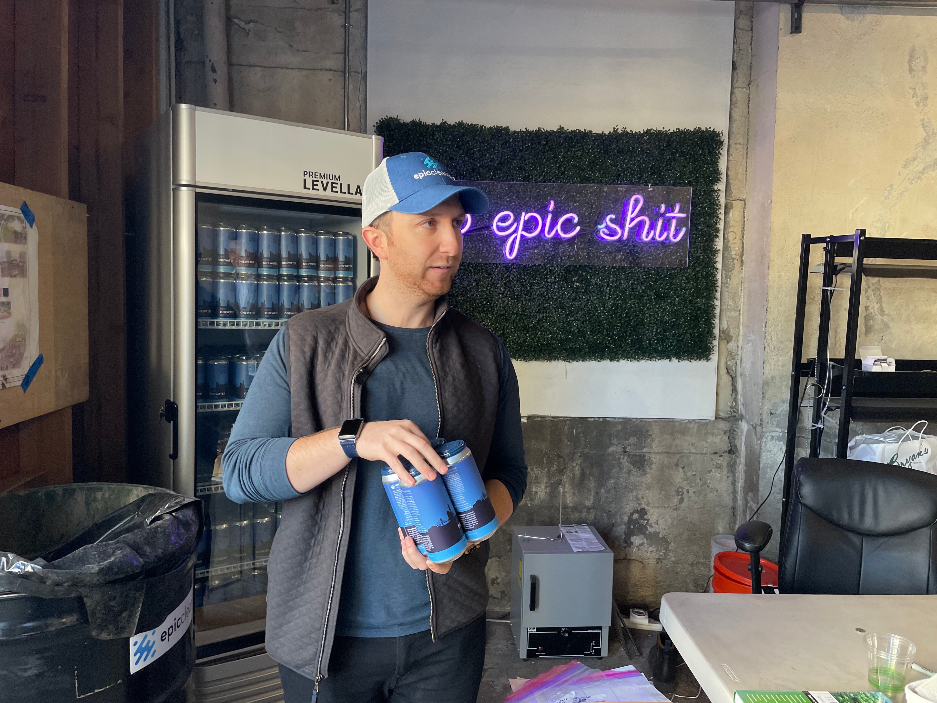 A man in a cap and a vest holds a four-pack of beer with a sign behind him that reads &quot;do epic shit.&quot;