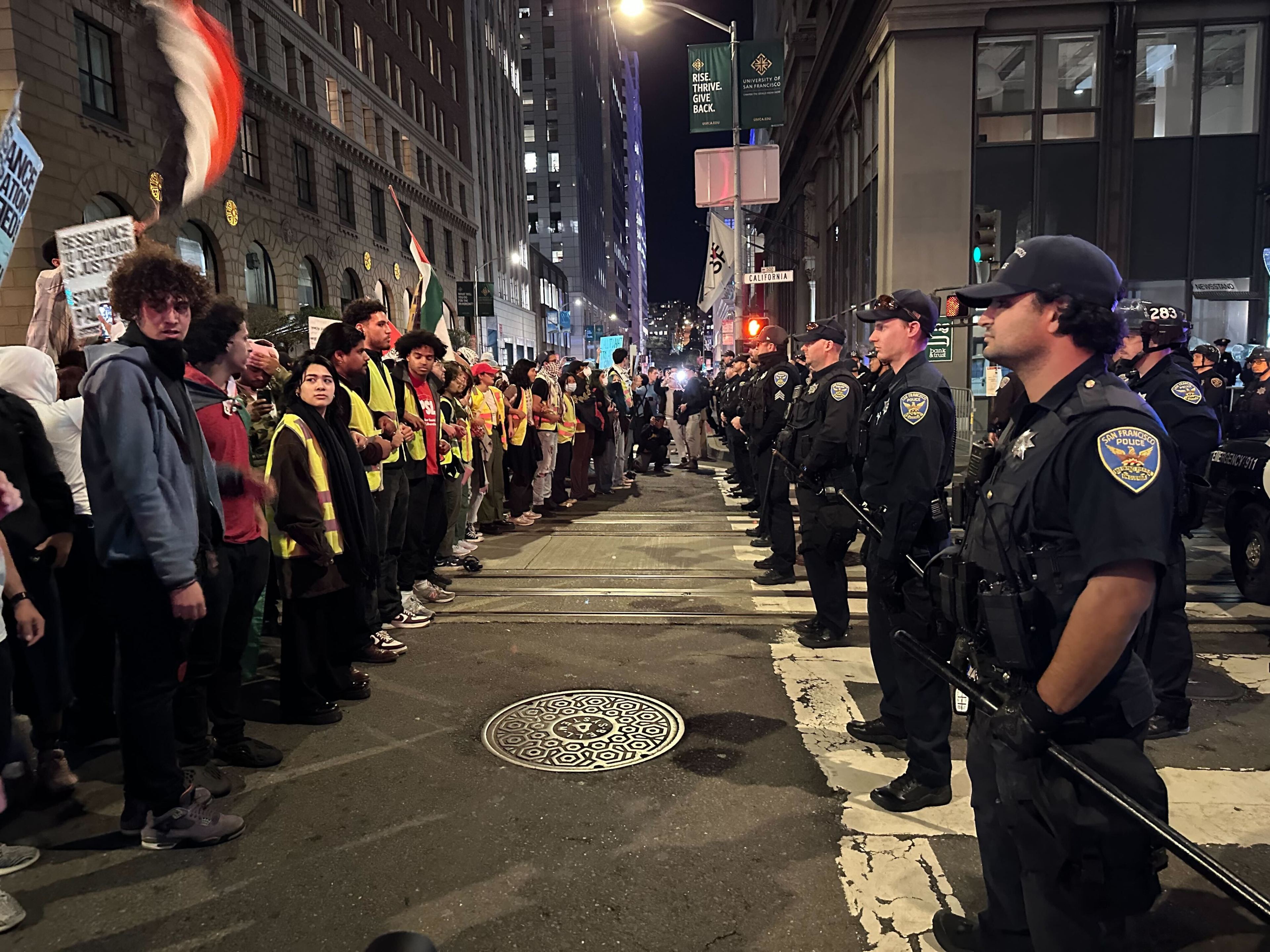 Protesters waving Palestinian flags stand in front of a row of San Francisco police officers on a city street.