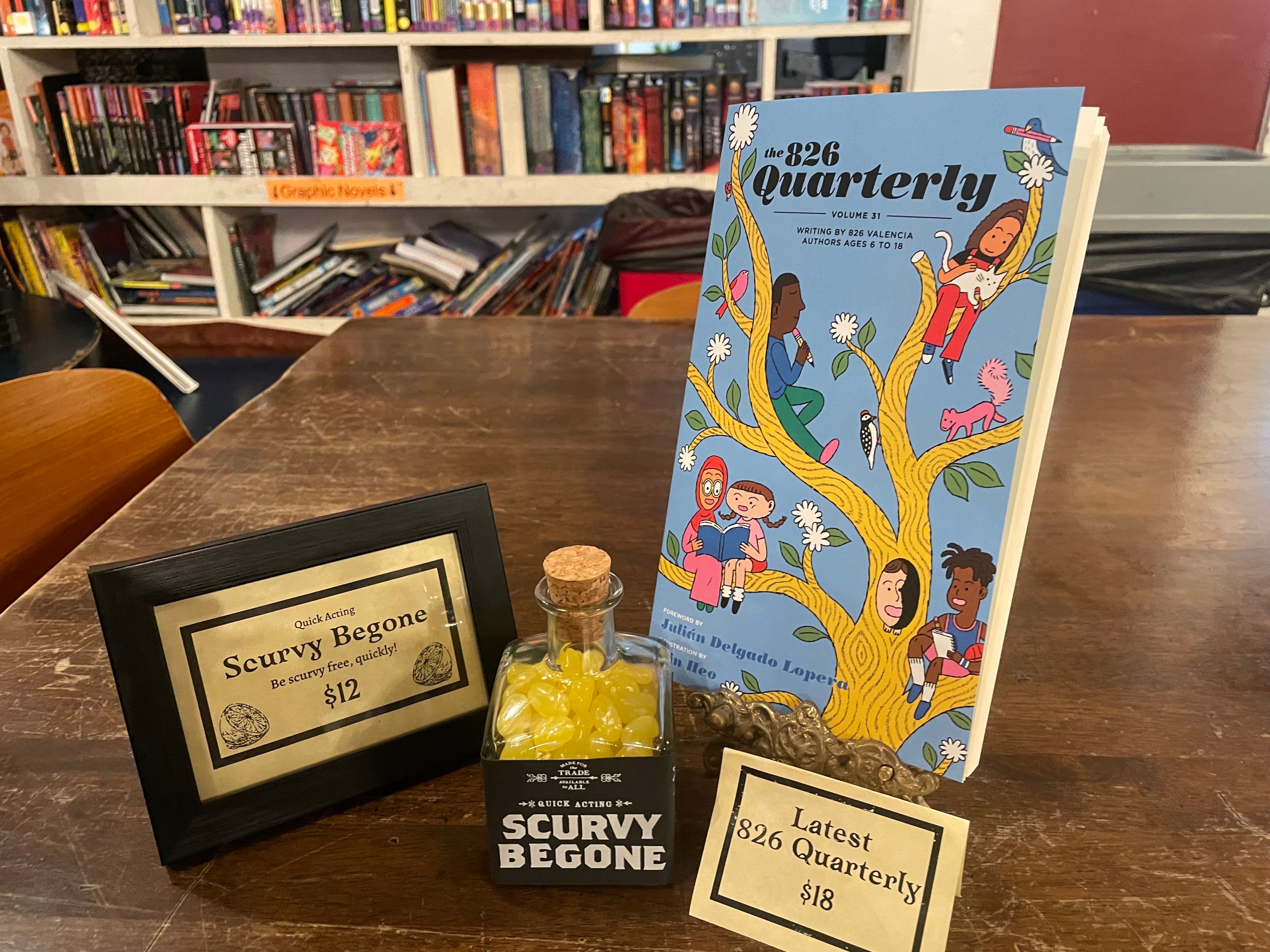 A &quot;Scurvy Begone&quot; bottle of candy and a children's literary journal sit on a a wooden table in a pirate-themed storefront.