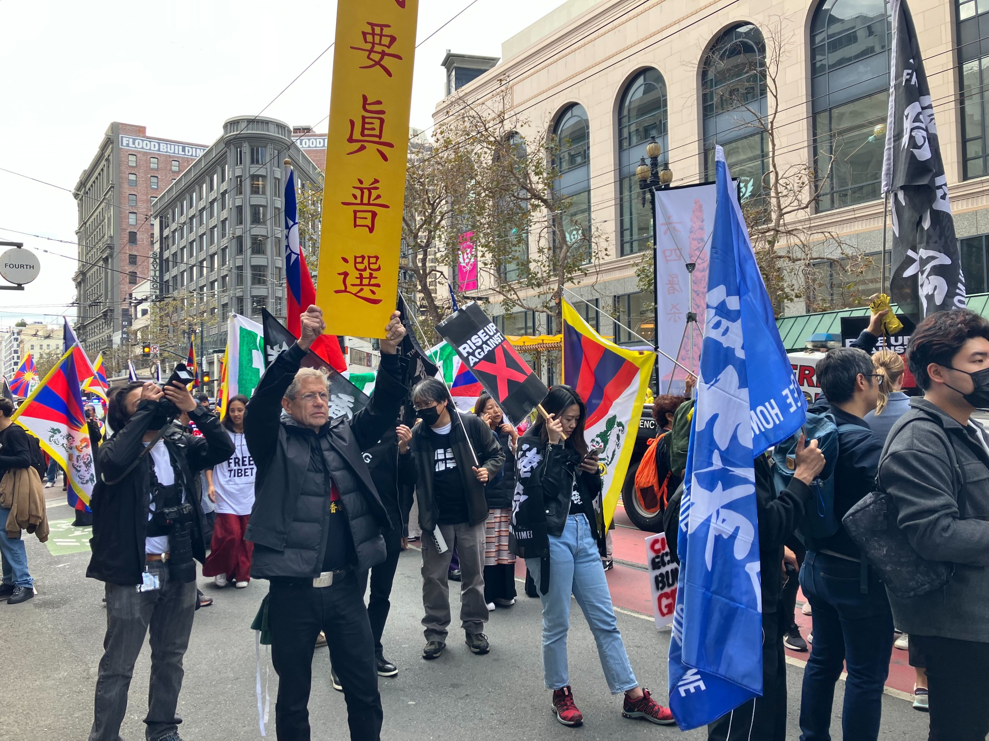 Protesters carrying colorful flags and signs in Chinese march down the street under cloudy skies.