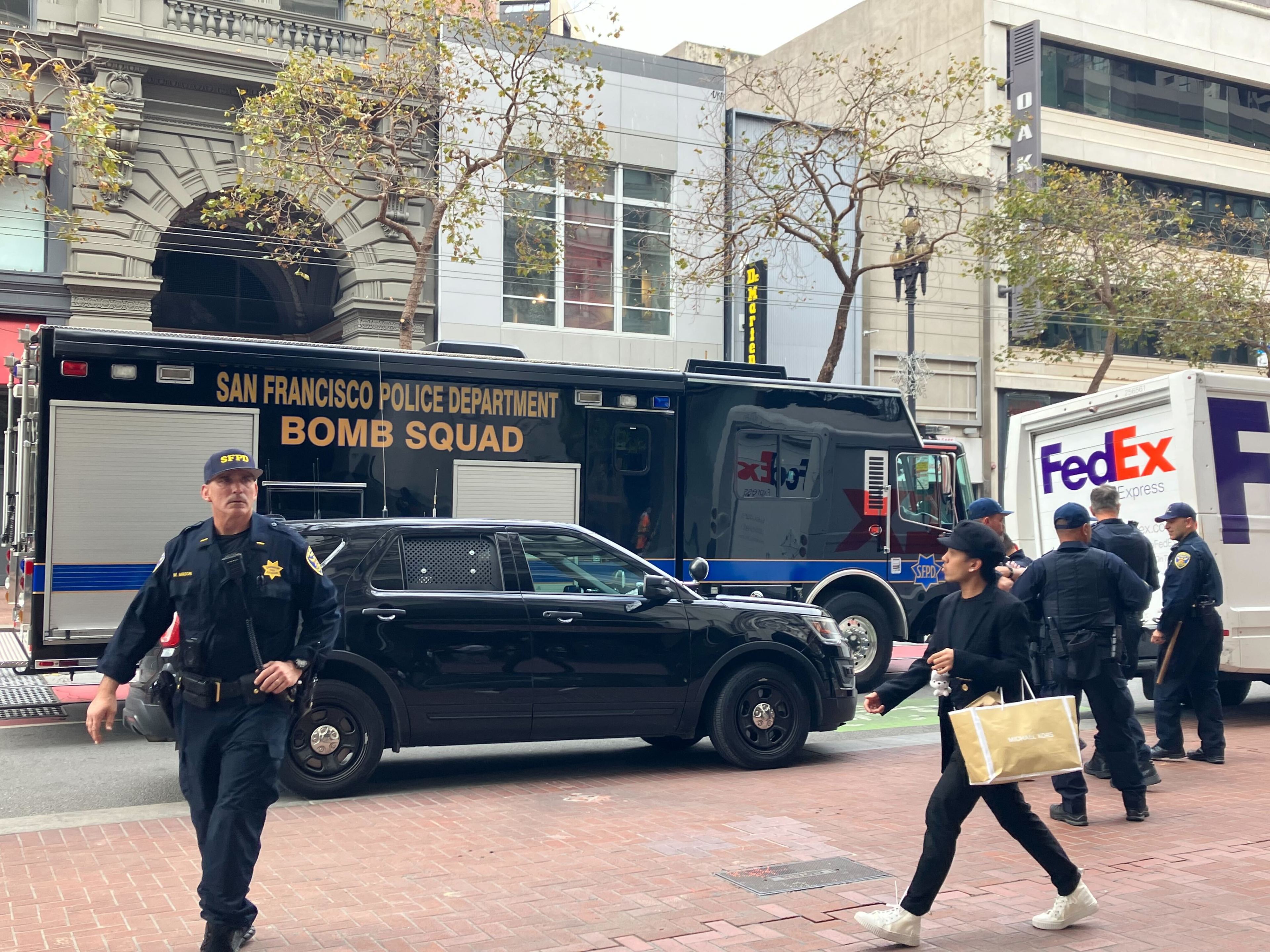Uniformed police officers are walking near a large dark bomb squad van on a busy street as pedestrians pass by.