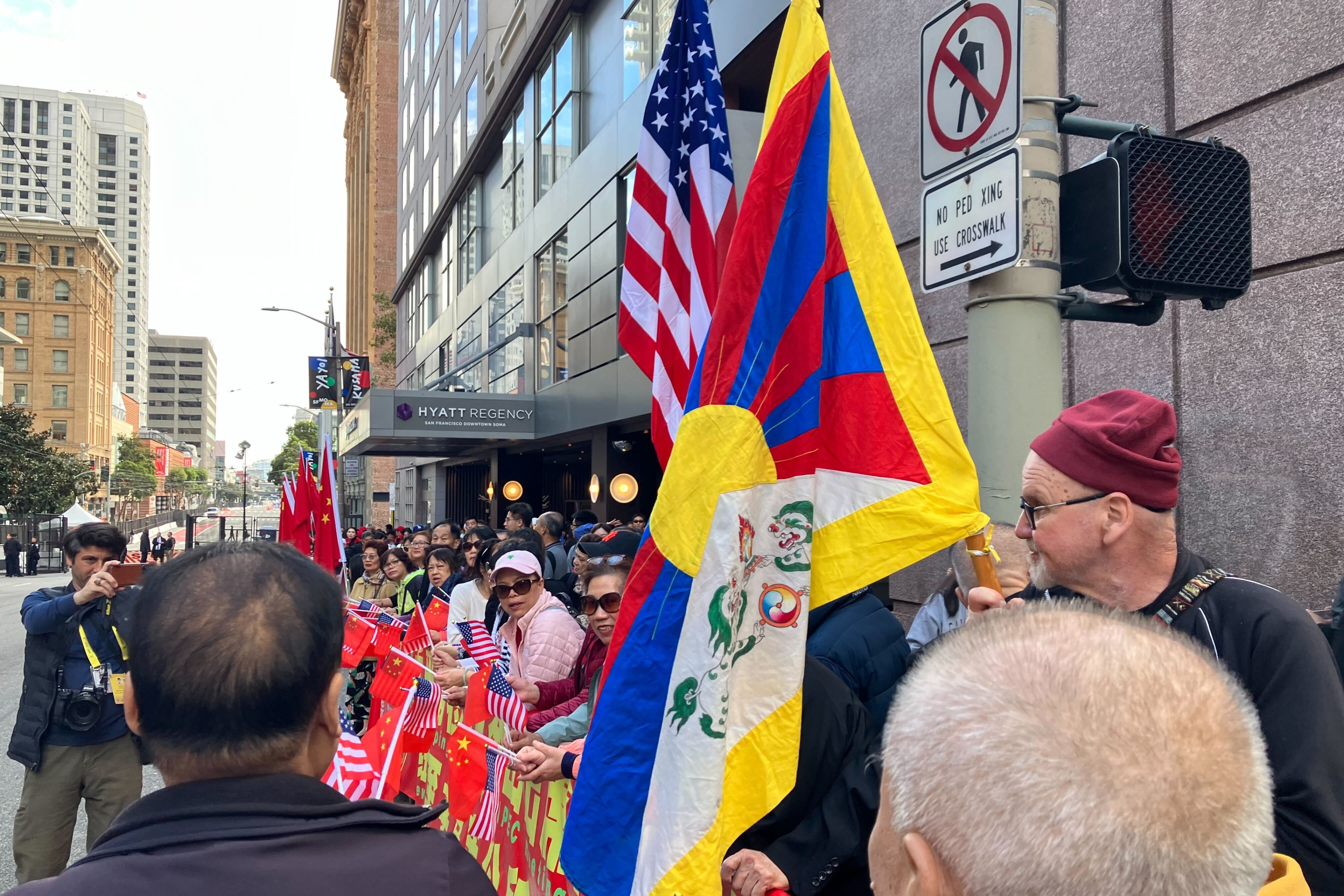 A long line of people stand along a city street holding American and Chinese flags and one man to the far right holds a Tibetan flag.